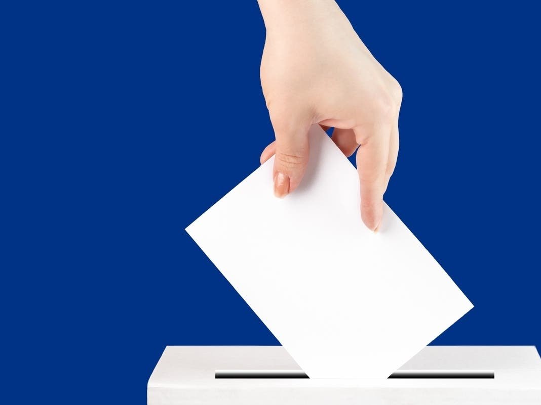 A hand inserting a white ballot paper into a white ballot box.
