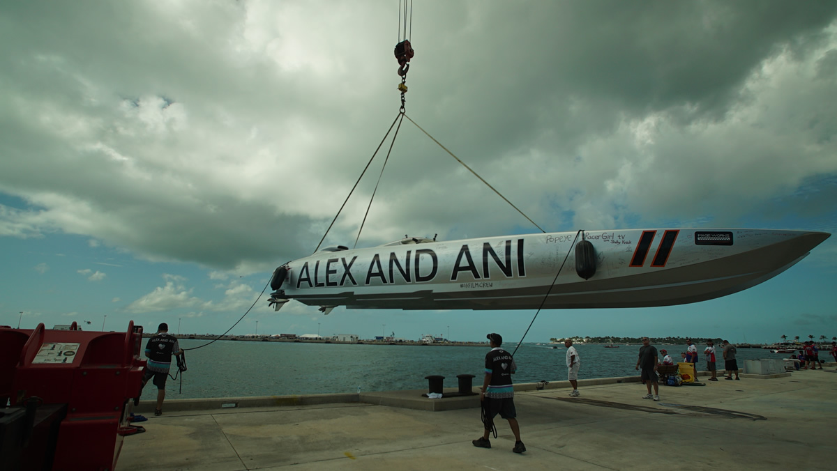 Offshore racing powerboat suspended by crane over a waterfront dock with crew standing nearby.