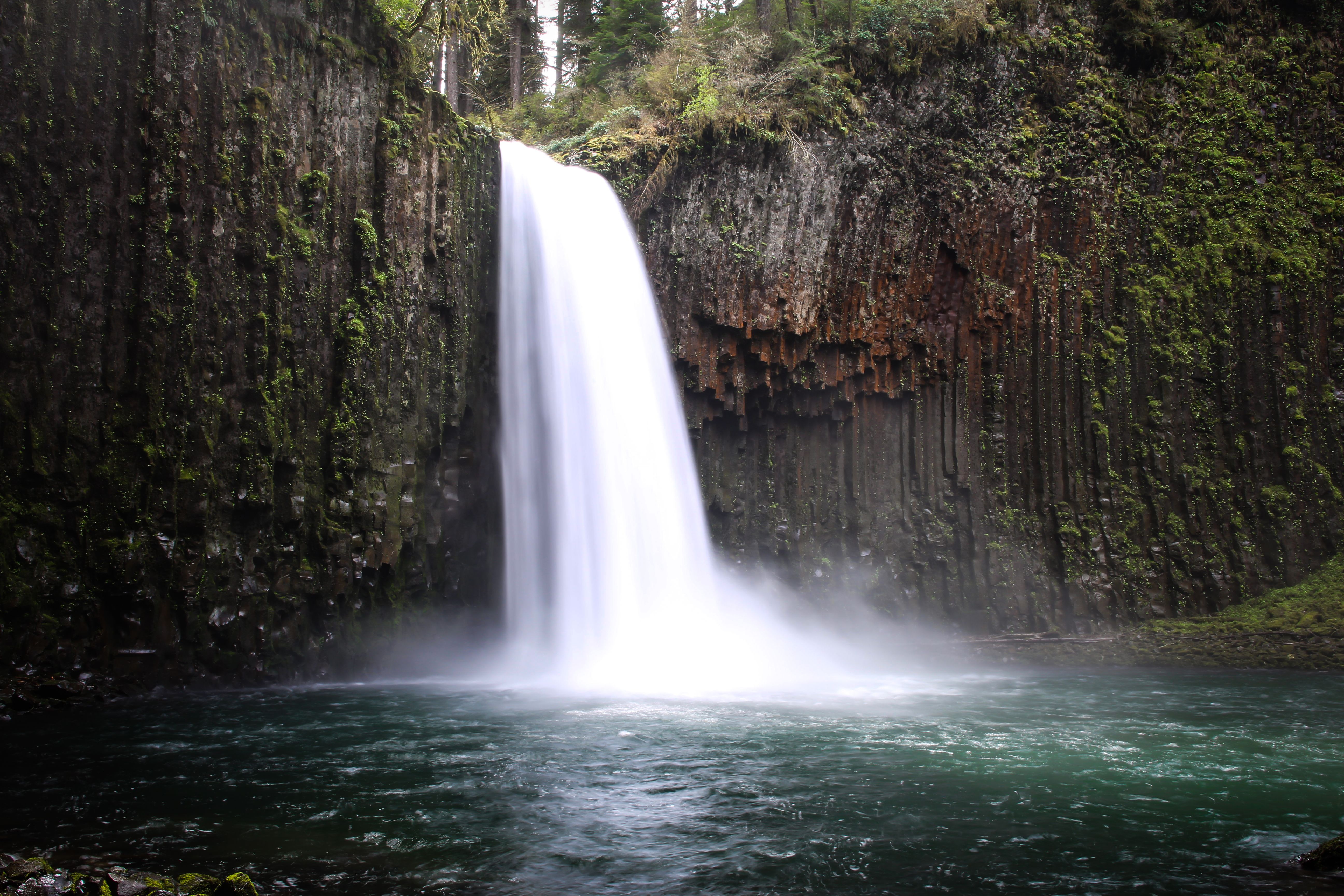 Tall waterfall cascading over moss-covered columnar basalt into a teal-green pool.