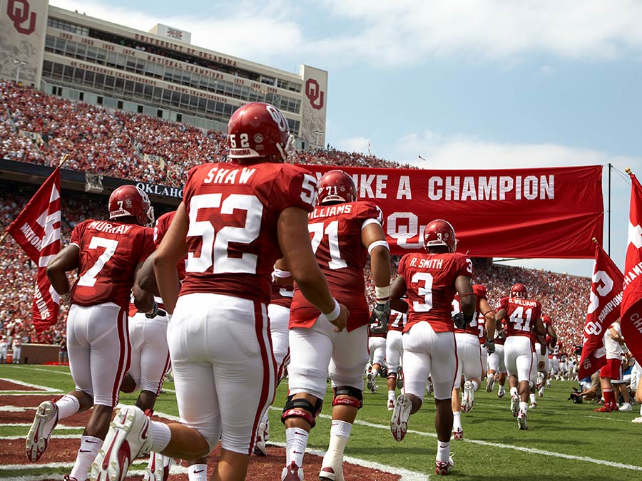 Football players in red uniforms run through a banner onto a field before a packed stadium crowd.