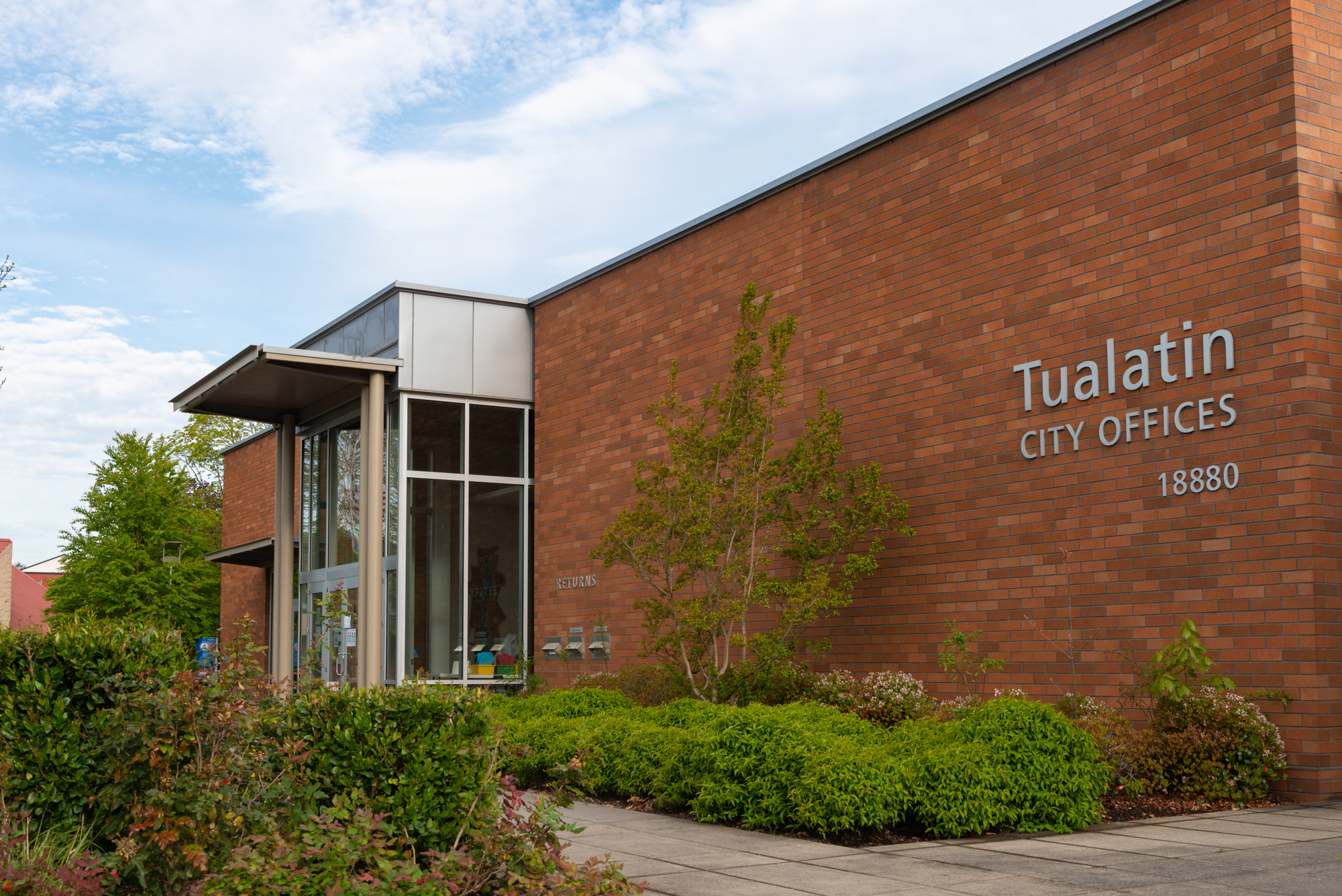 Red brick city offices building with glass entryway and landscaped shrubs.