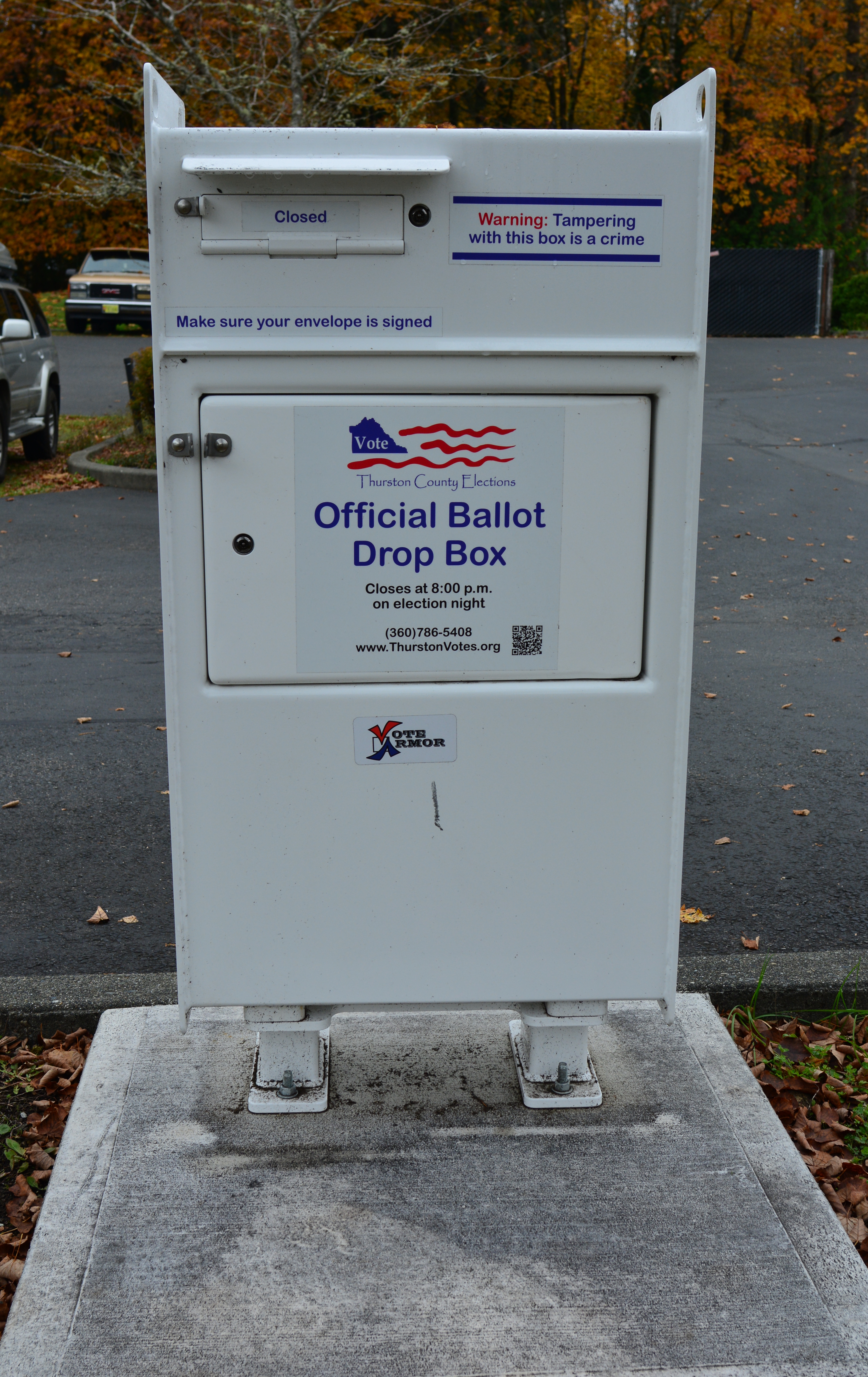 Close-up of a white official ballot drop box with signage and instructions in a parking lot.