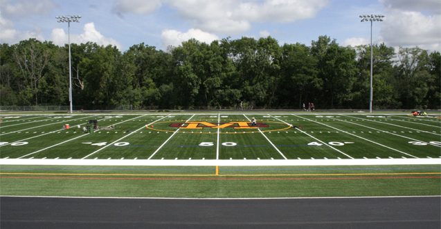 Aerial view of an artificial turf football field with a gold and black midfield logo.
