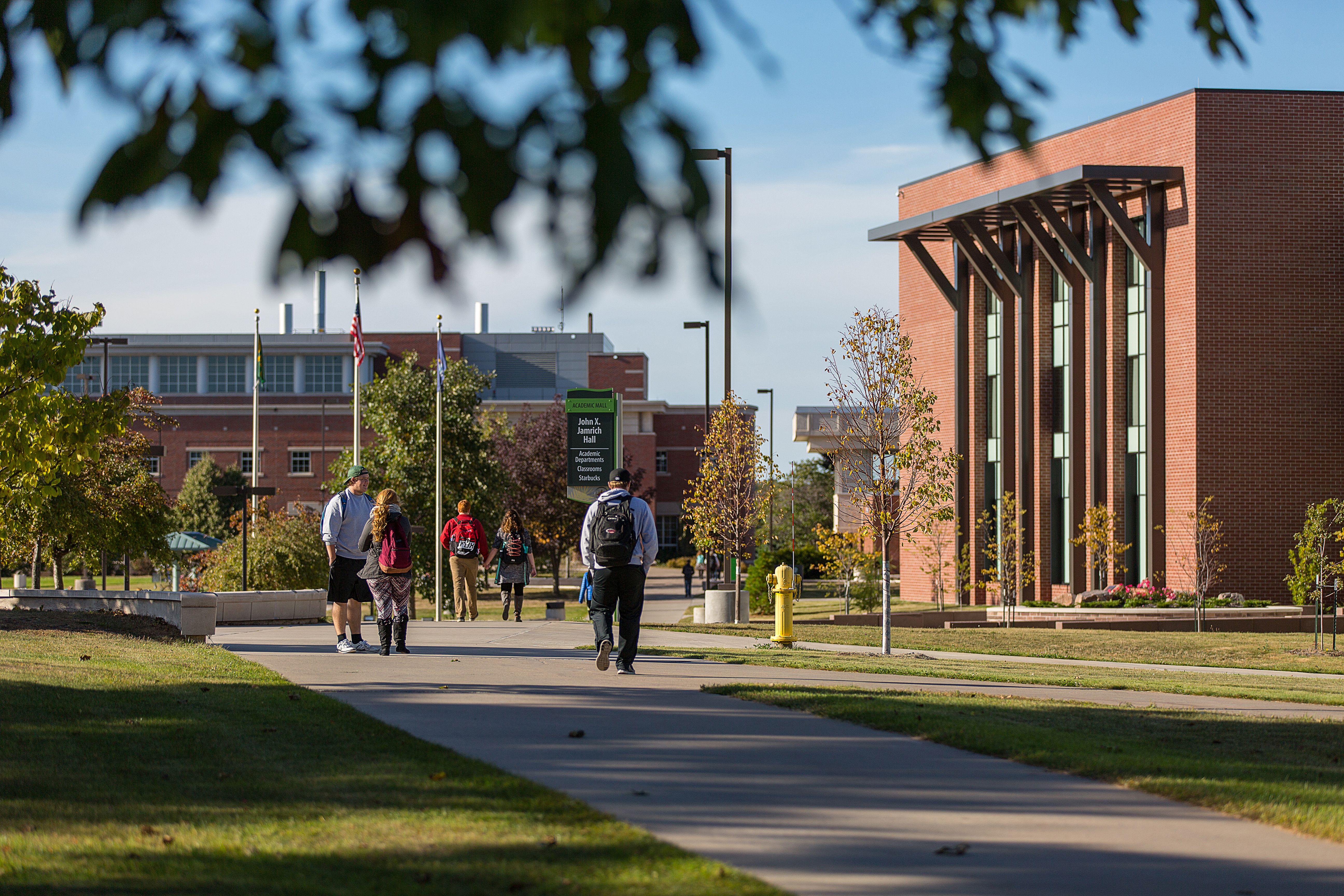 Students walk along a brick campus pathway flanked by academic buildings and flagpoles on a sunny day.