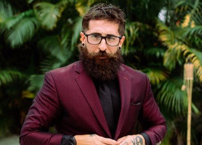 Bearded man in burgundy blazer and black turtleneck standing before tropical foliage.