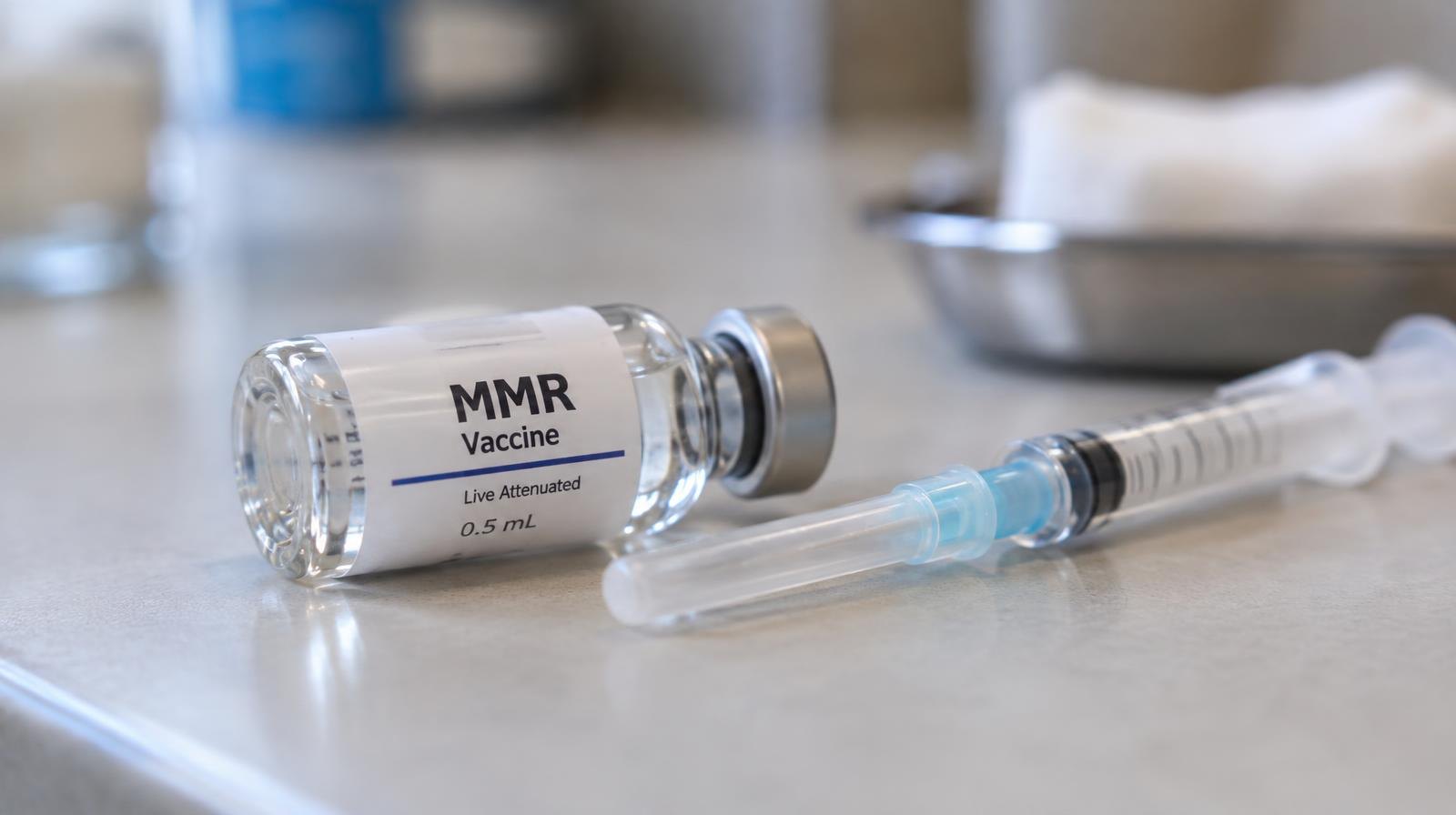 A glass vaccine vial lying on its side next to a capped syringe on a clean surface.