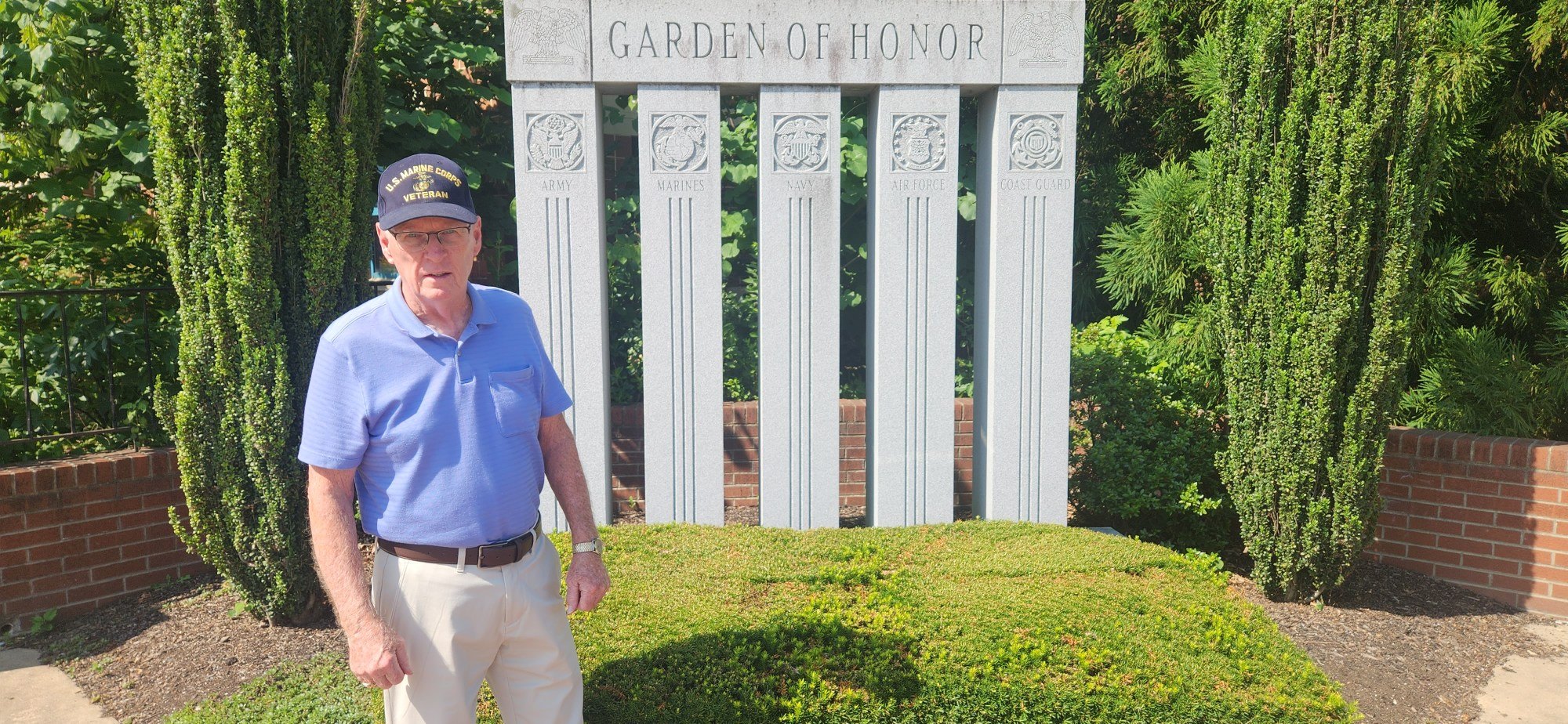 Elderly man in blue polo shirt and cap stands before a white stone Garden of Honor monument.