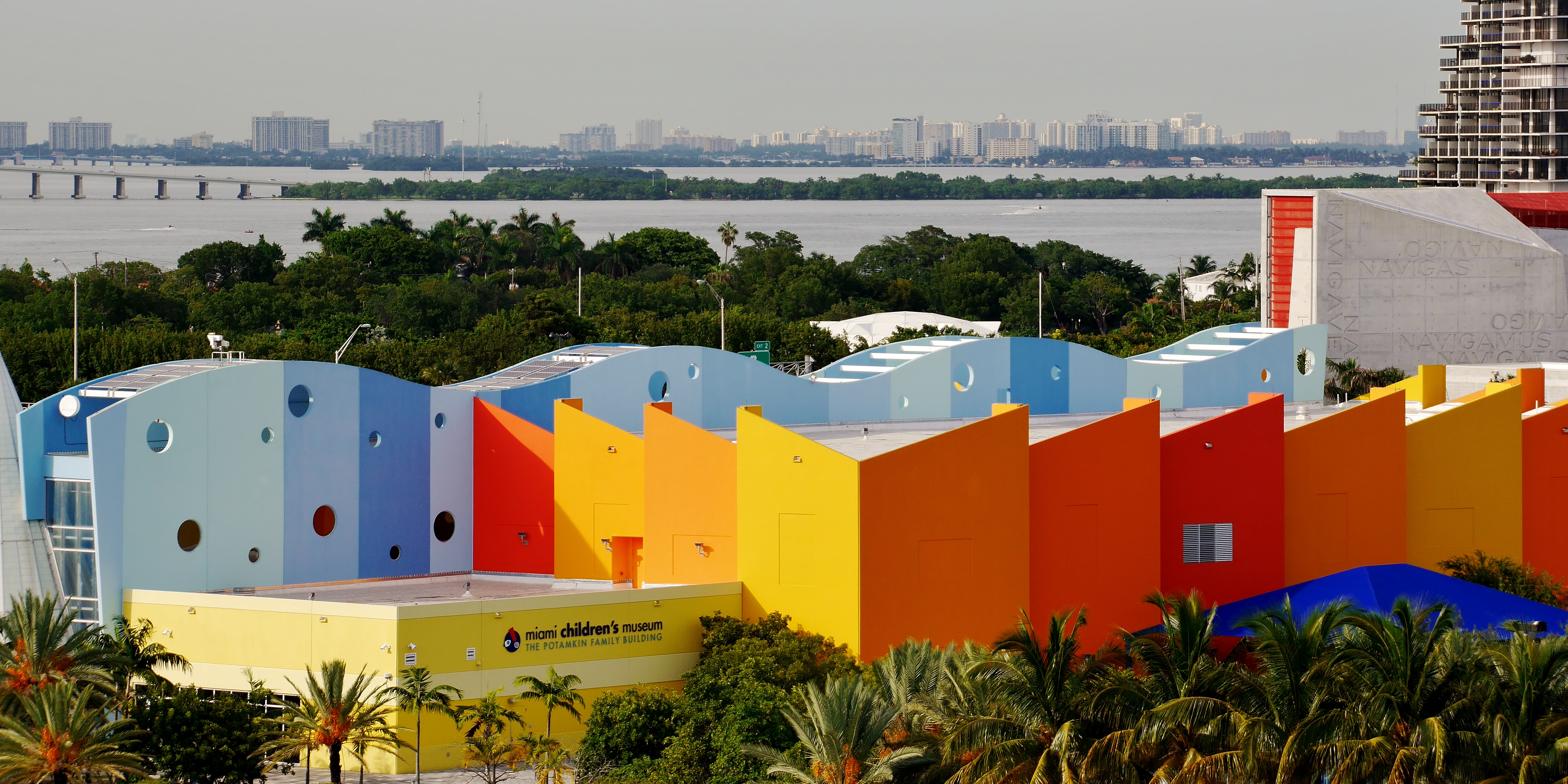 Colorful building exterior with wave-shaped roofline, palm trees, and a waterway in the foreground.