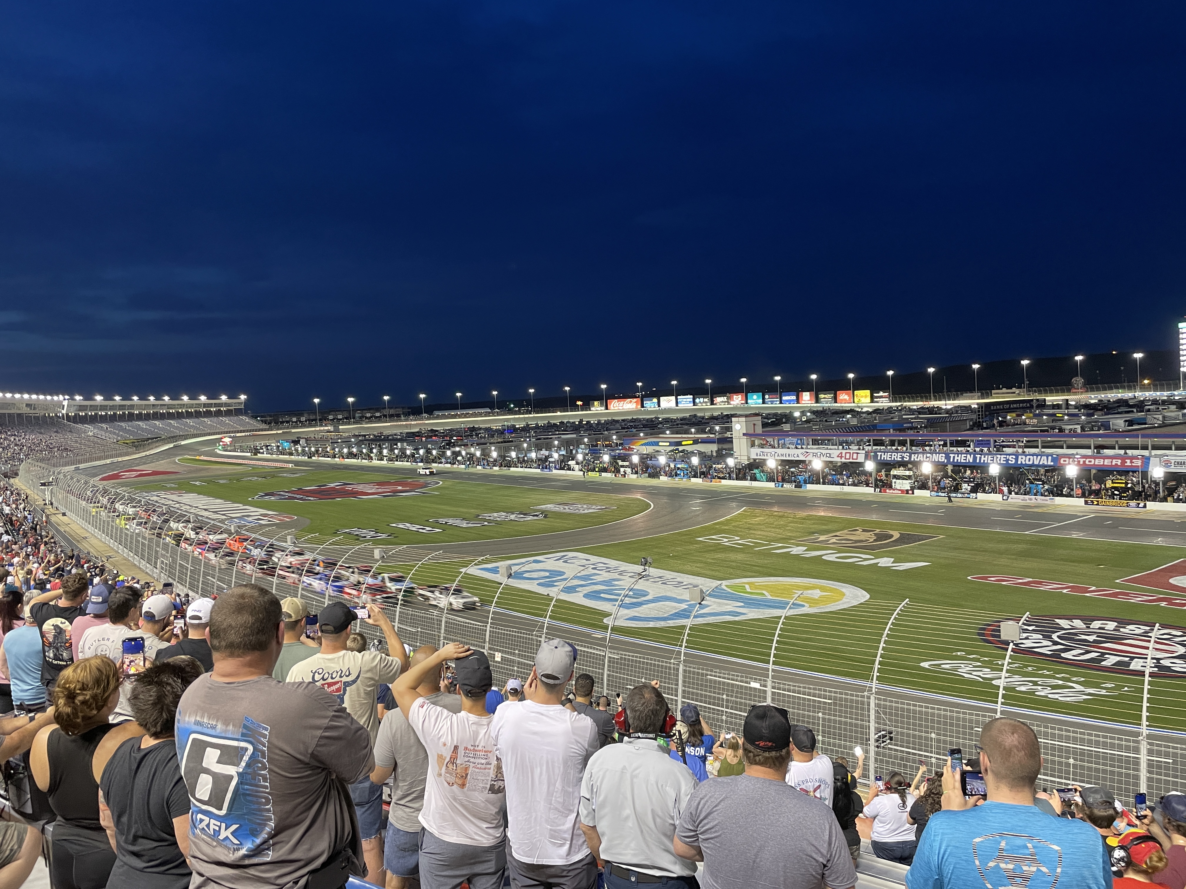 Nighttime NASCAR race at Charlotte Motor Speedway with packed grandstands and cars on track.
