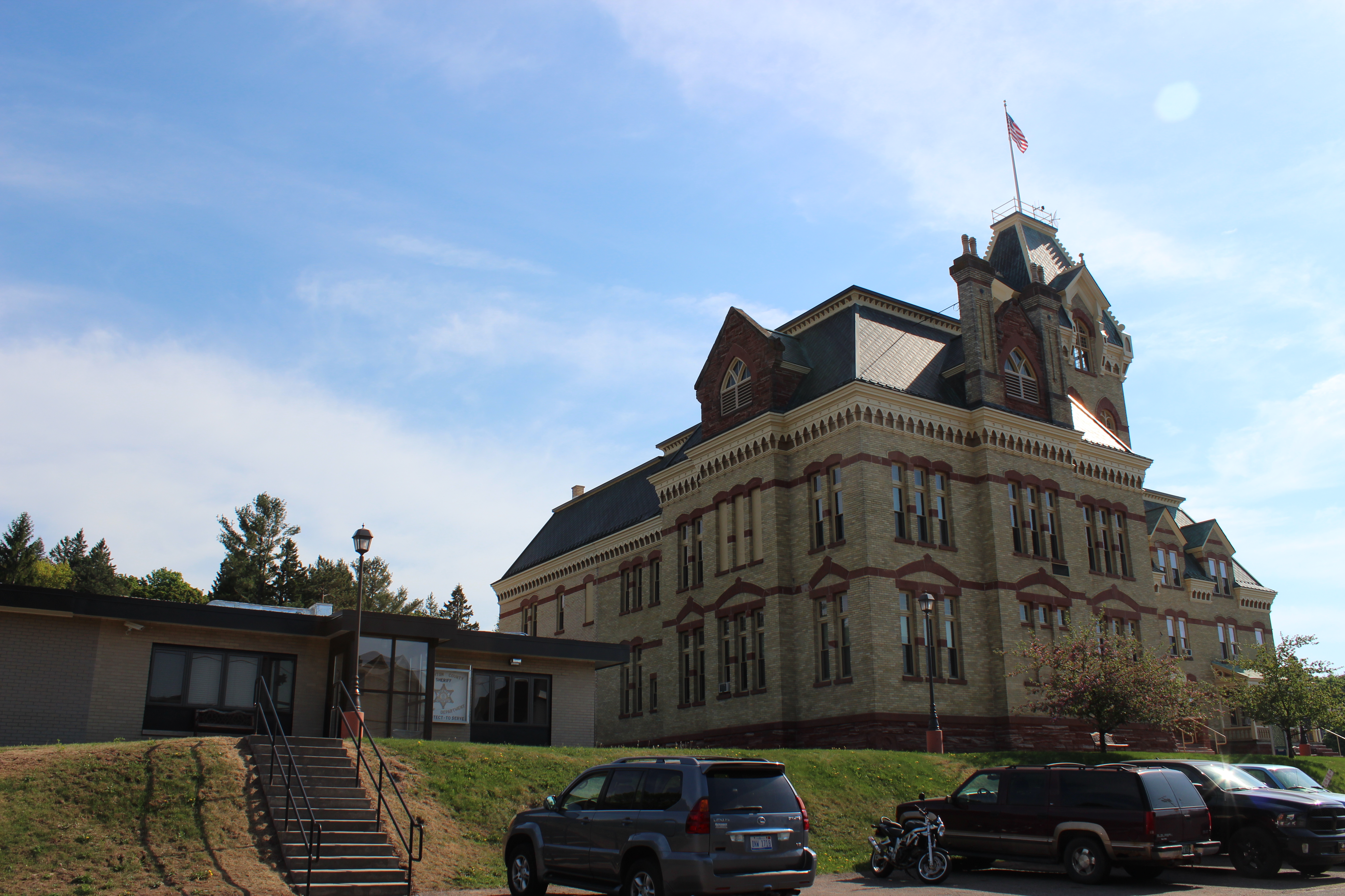 Historic stone courthouse with a central tower and flag, next to a smaller modern building.