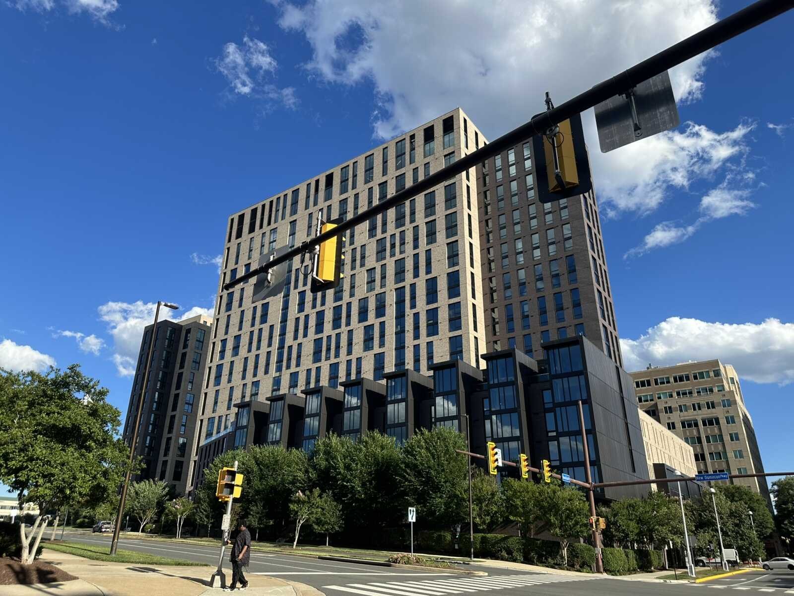 A modern high-rise apartment building with beige and dark facade at a street intersection with traffic lights and pedestrians.