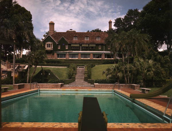 Historic mansion with red tile roof and rectangular pool surrounded by palm trees.