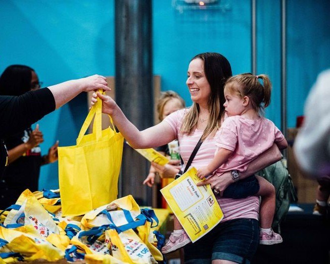 Smiling woman holding a toddler receives a yellow tote bag across a table.