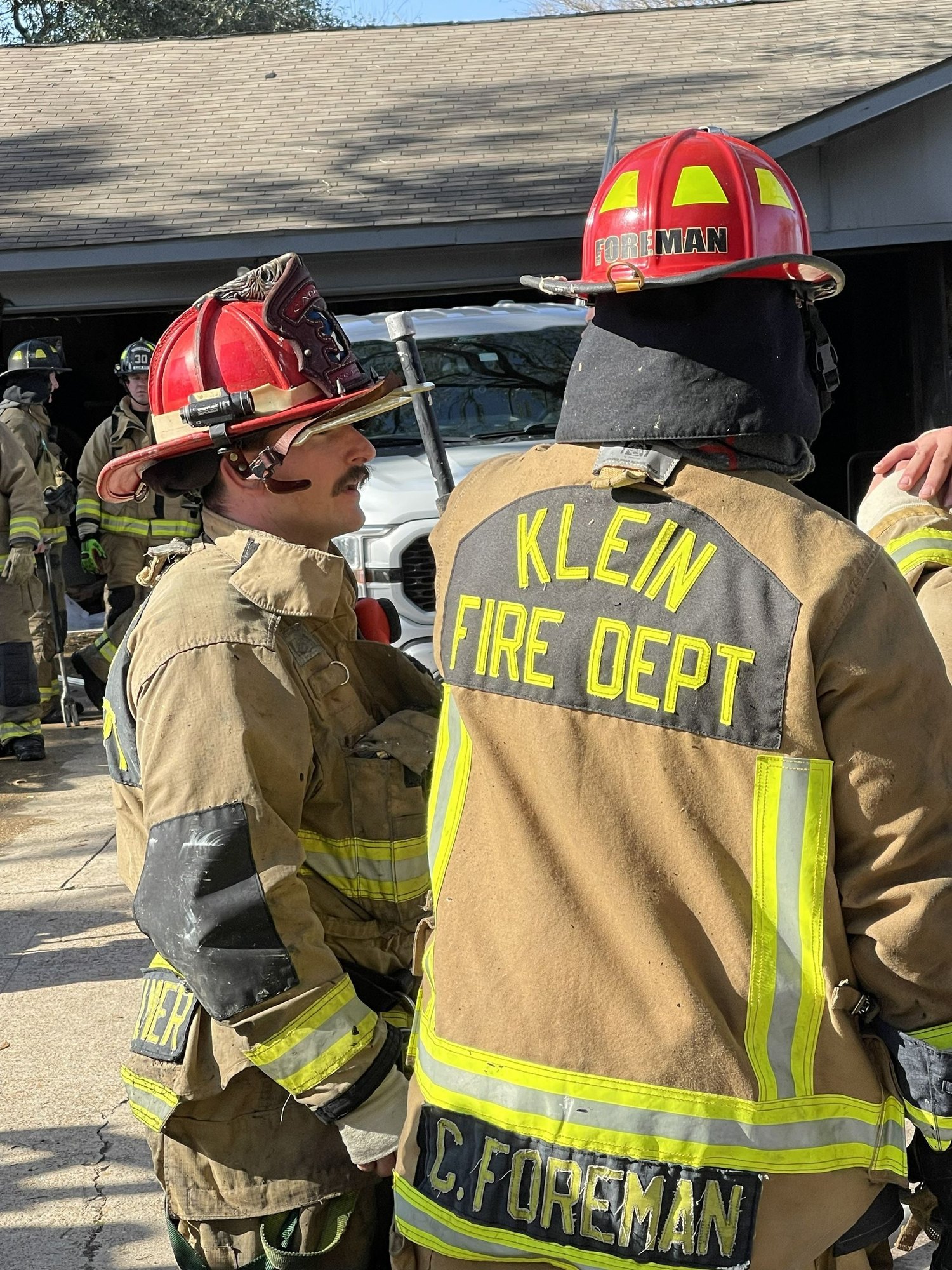 Two firefighters in turnout gear with red helmets face each other near a structure.