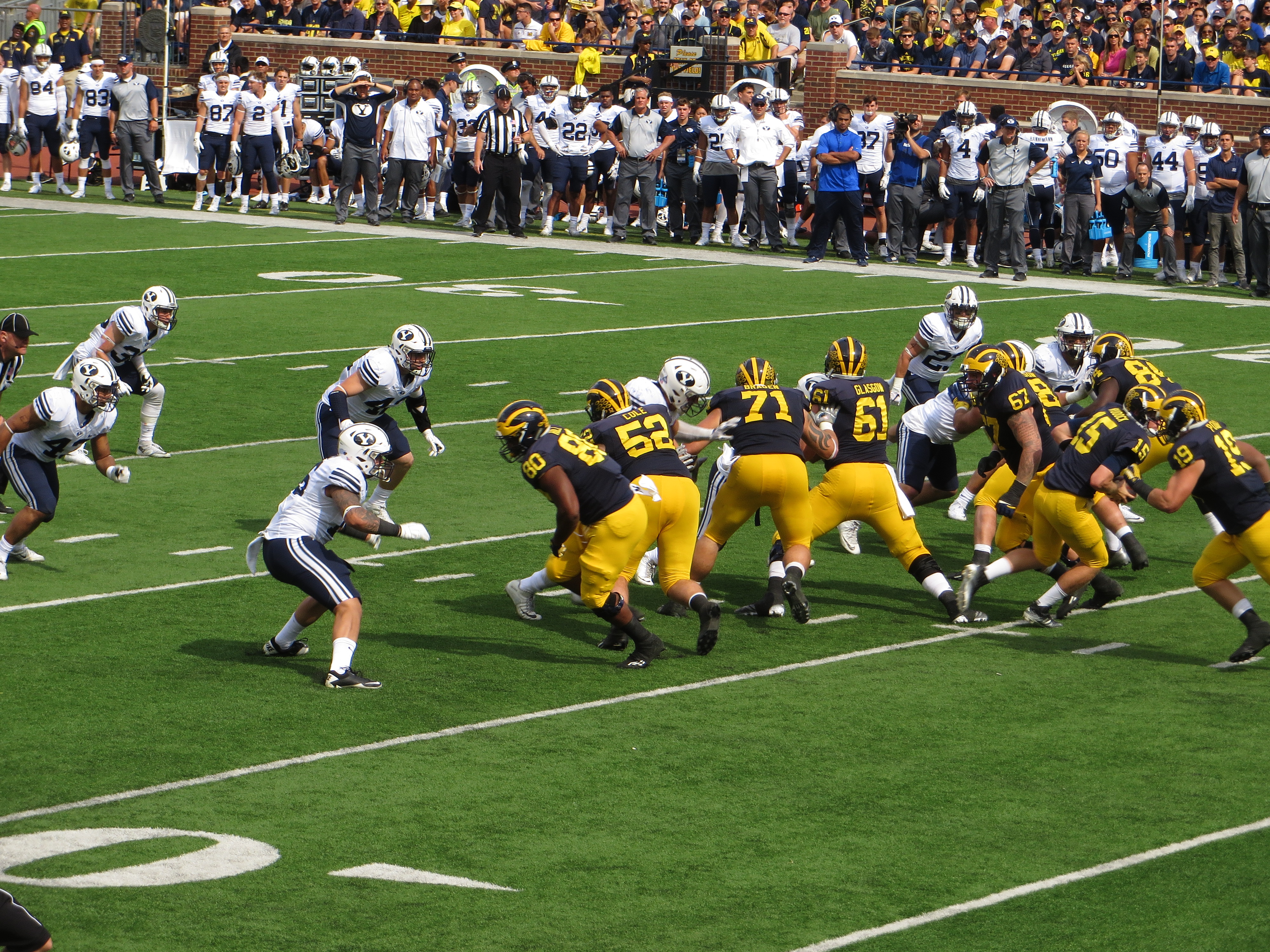 Michigan offensive linemen in maize and blue uniforms blocking against defenders in white at Michigan Stadium.