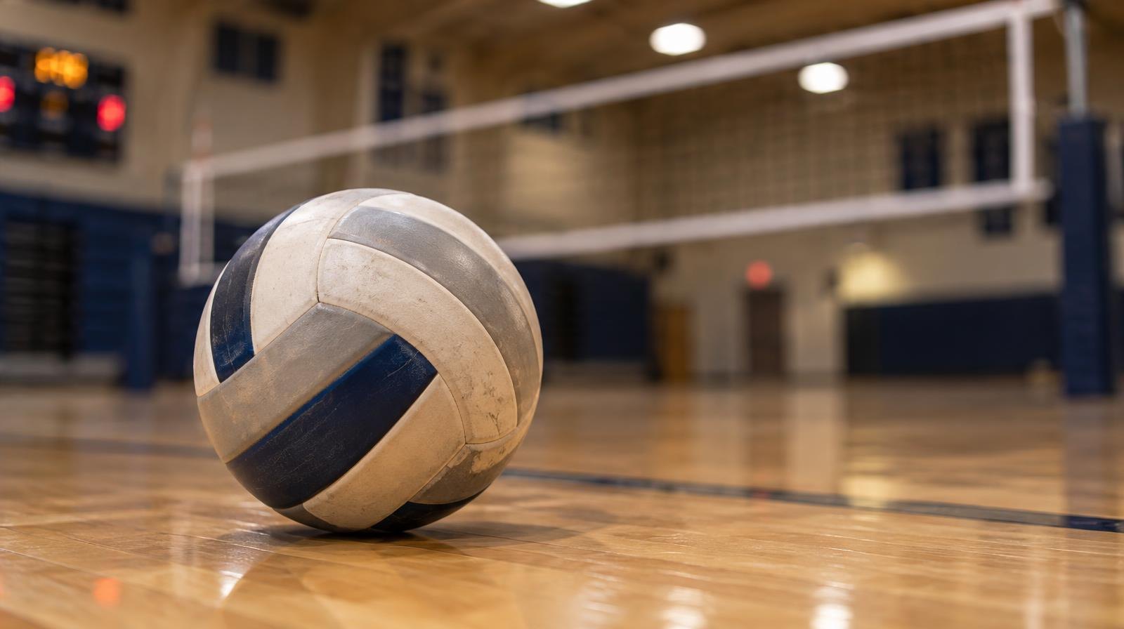 A volleyball resting on a hardwood gymnasium floor with a net in soft focus behind it.