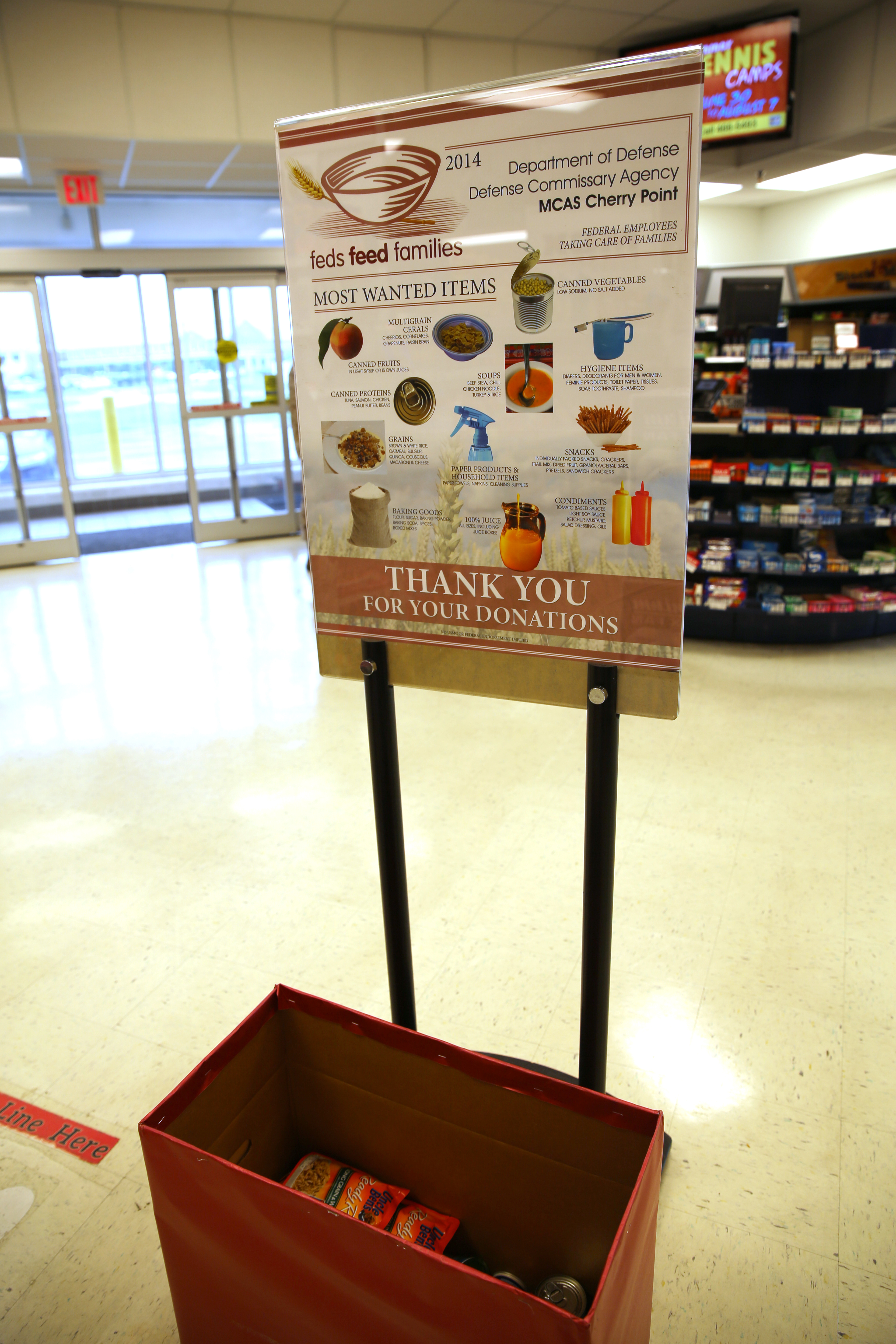 A red donation box filled with canned goods in front of a food drive sign.