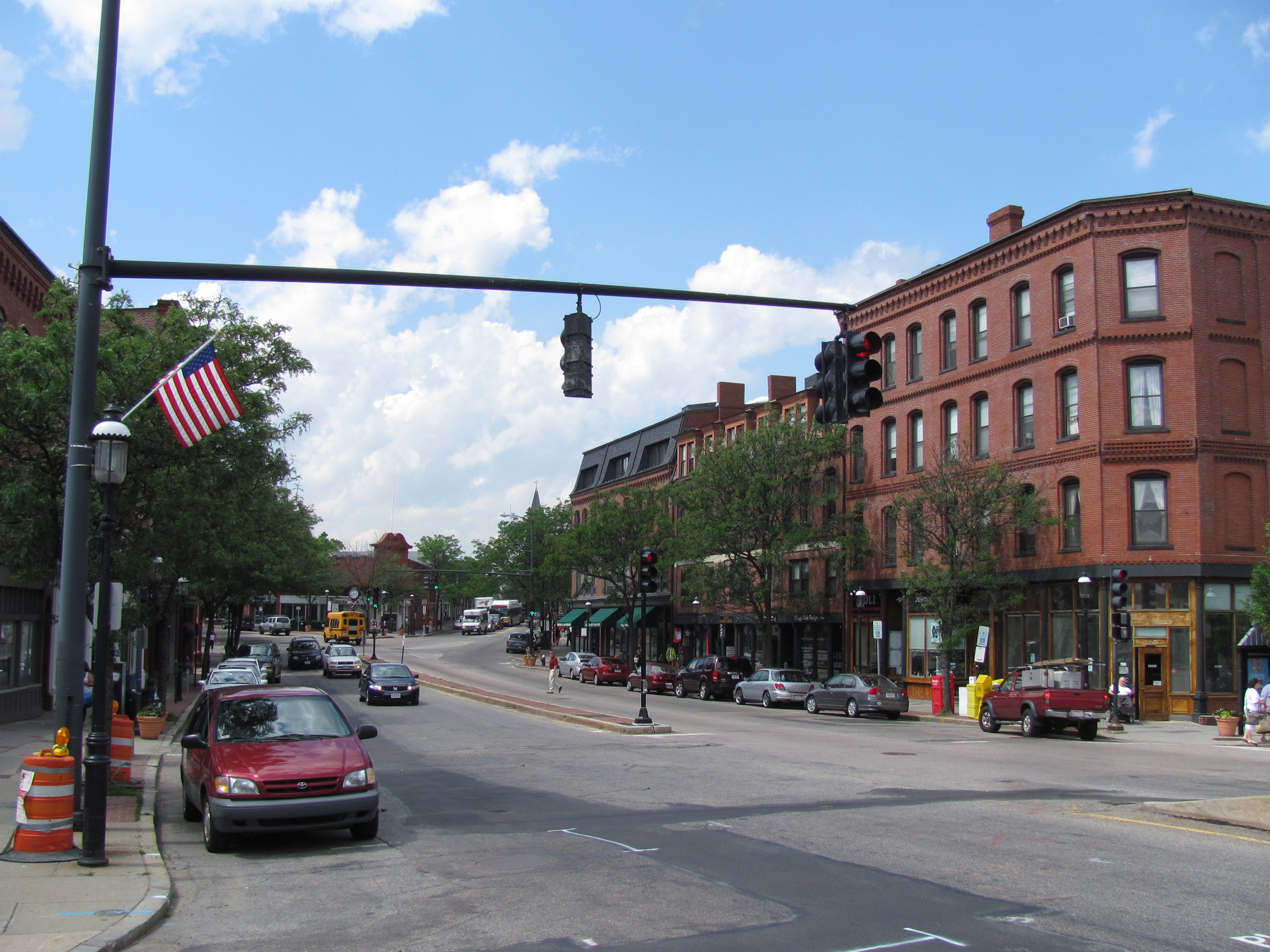 Intersection of two streets in Brookline Village with red-brick buildings, traffic signals, and an American flag.