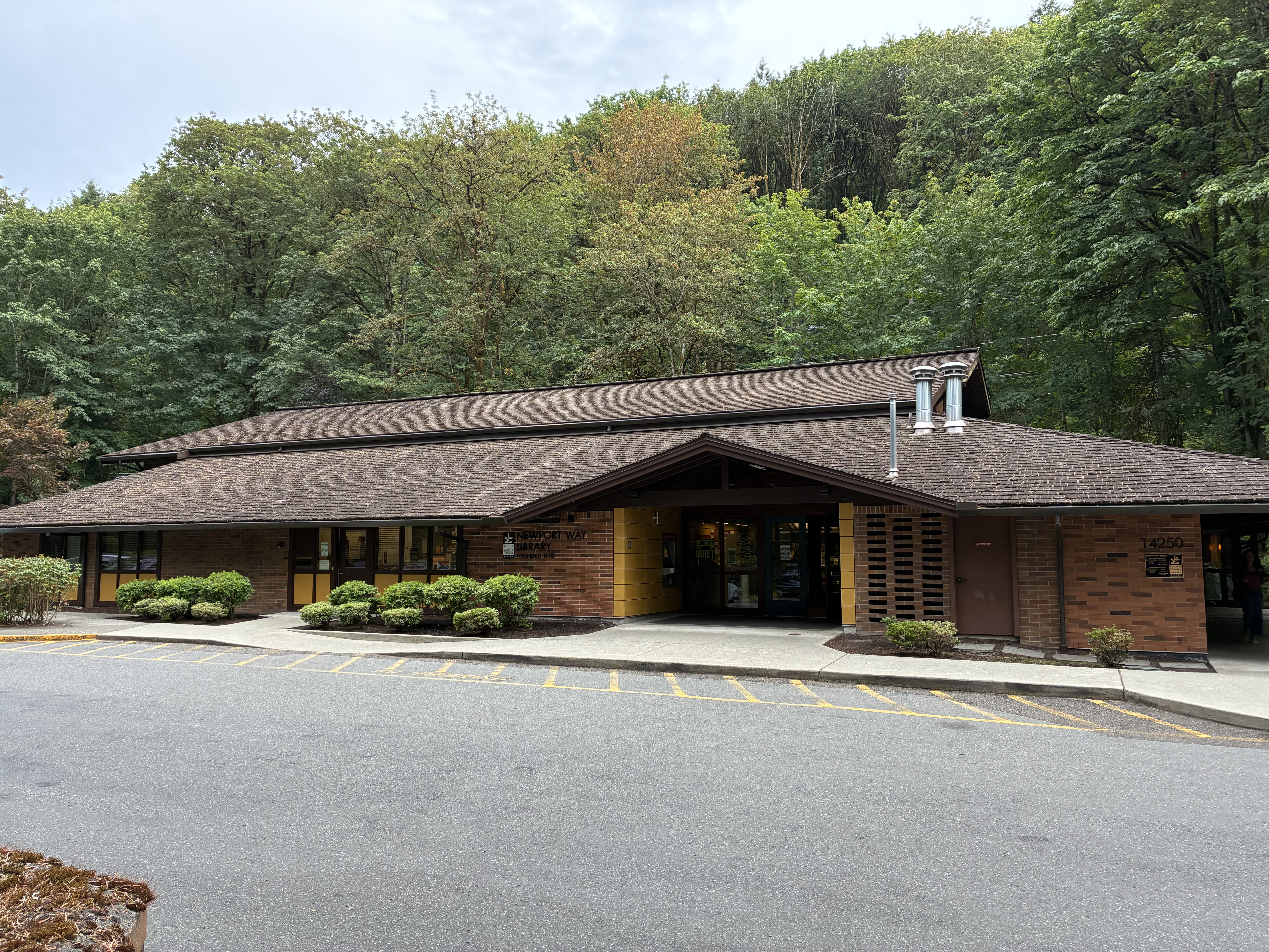 Single-story brick library building with a shingled roof surrounded by tall green trees.