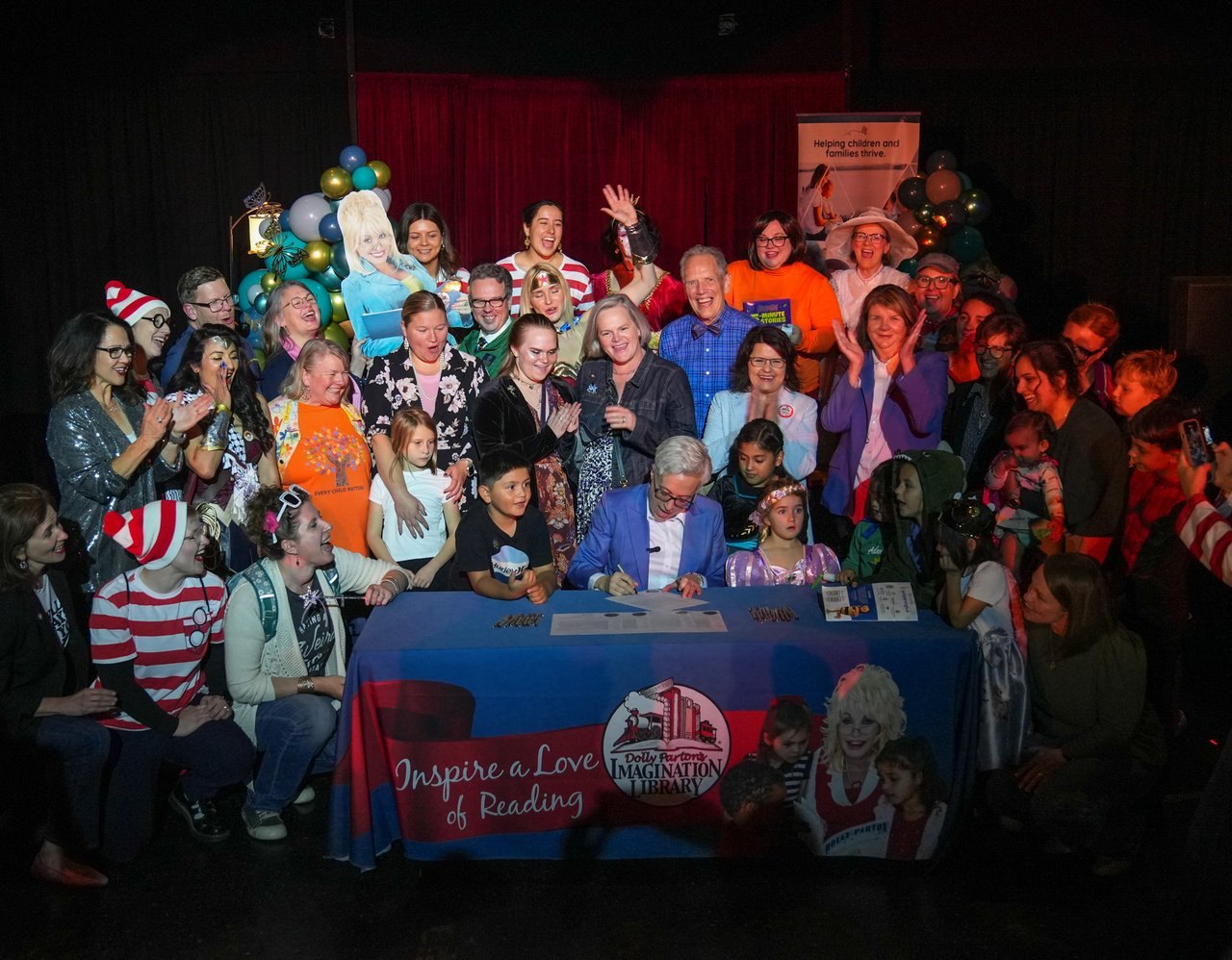 A crowd gathers around a signing table with an Imagination Library banner and a Dolly Parton cardboard cutout.