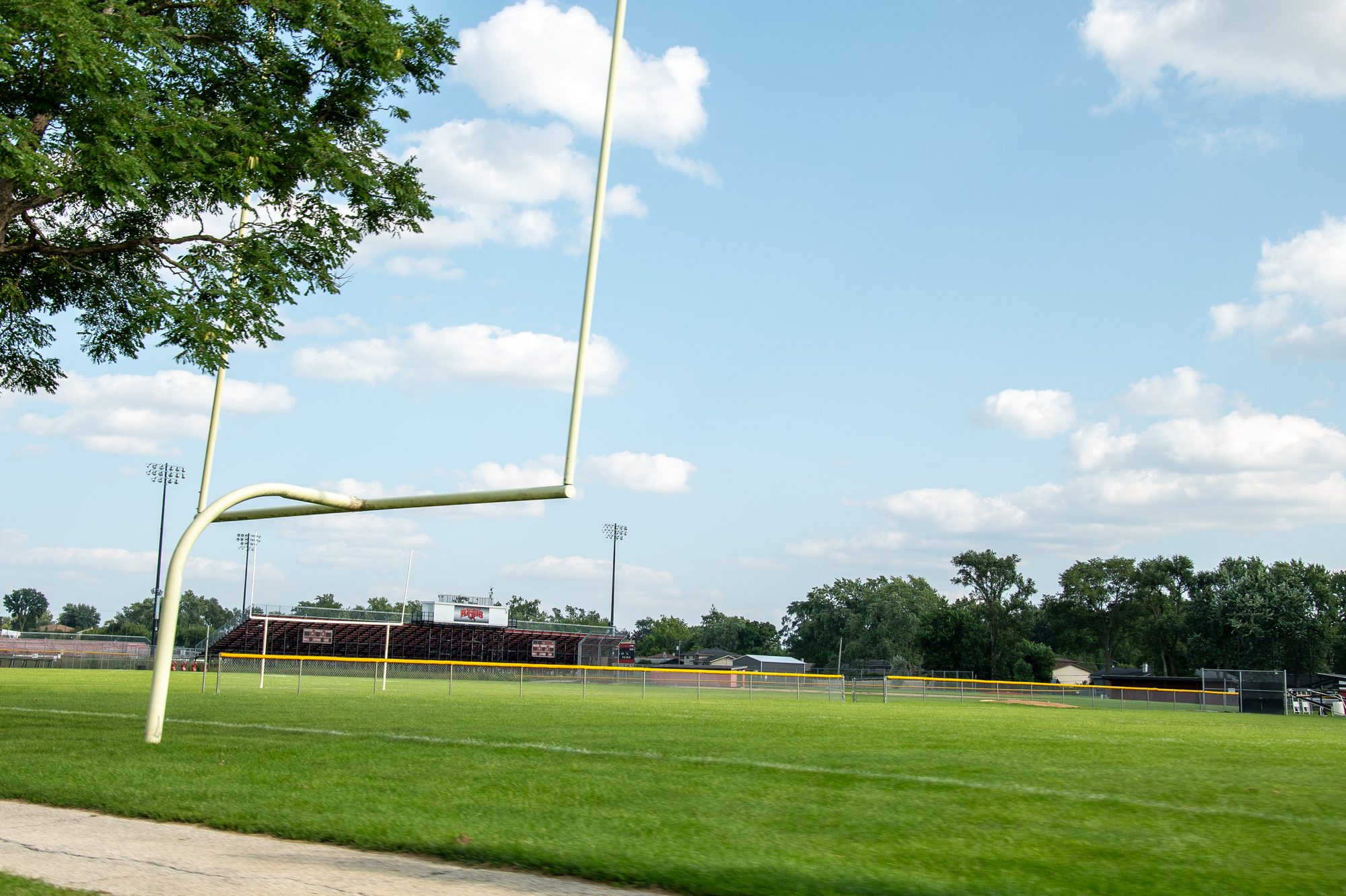 Yellow football goalpost on a grass field with stadium bleachers and scoreboard in background.