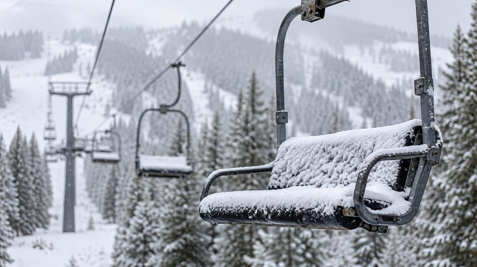 Snow-covered chairlift chair with pine trees and mountain slopes behind it.