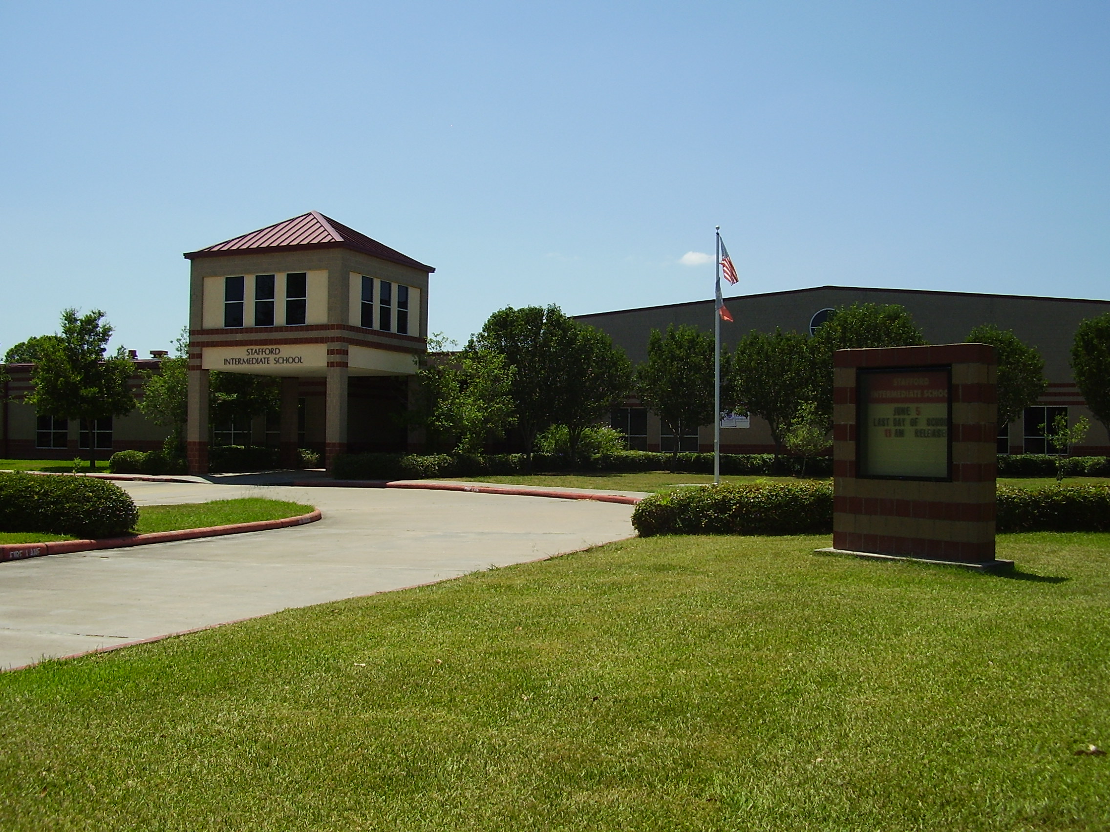 Brick school building entrance with red roof, flagpole, and manicured lawn.