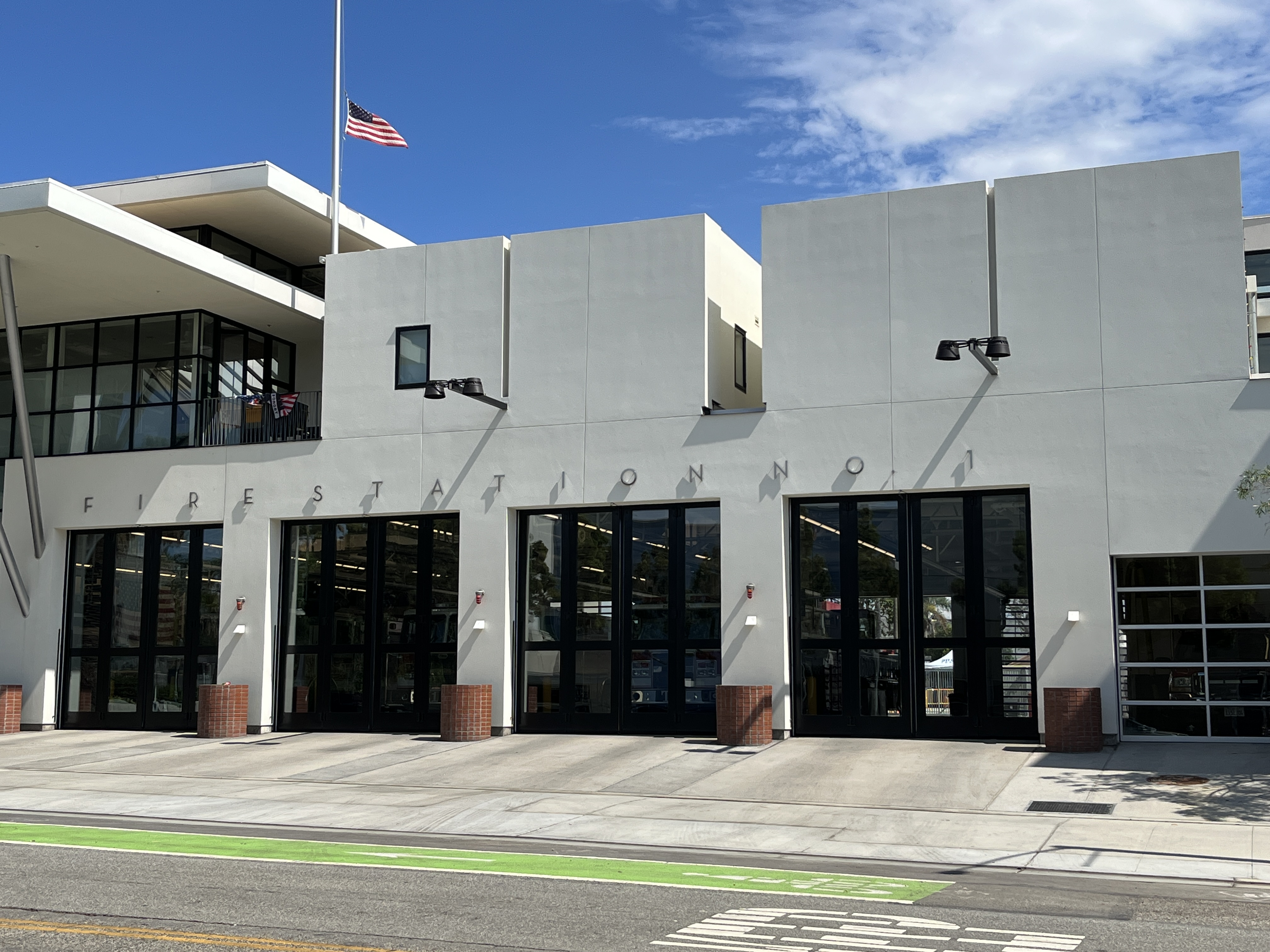 Exterior of a modern white fire station with glass bay doors and an American flag on a flagpole.
