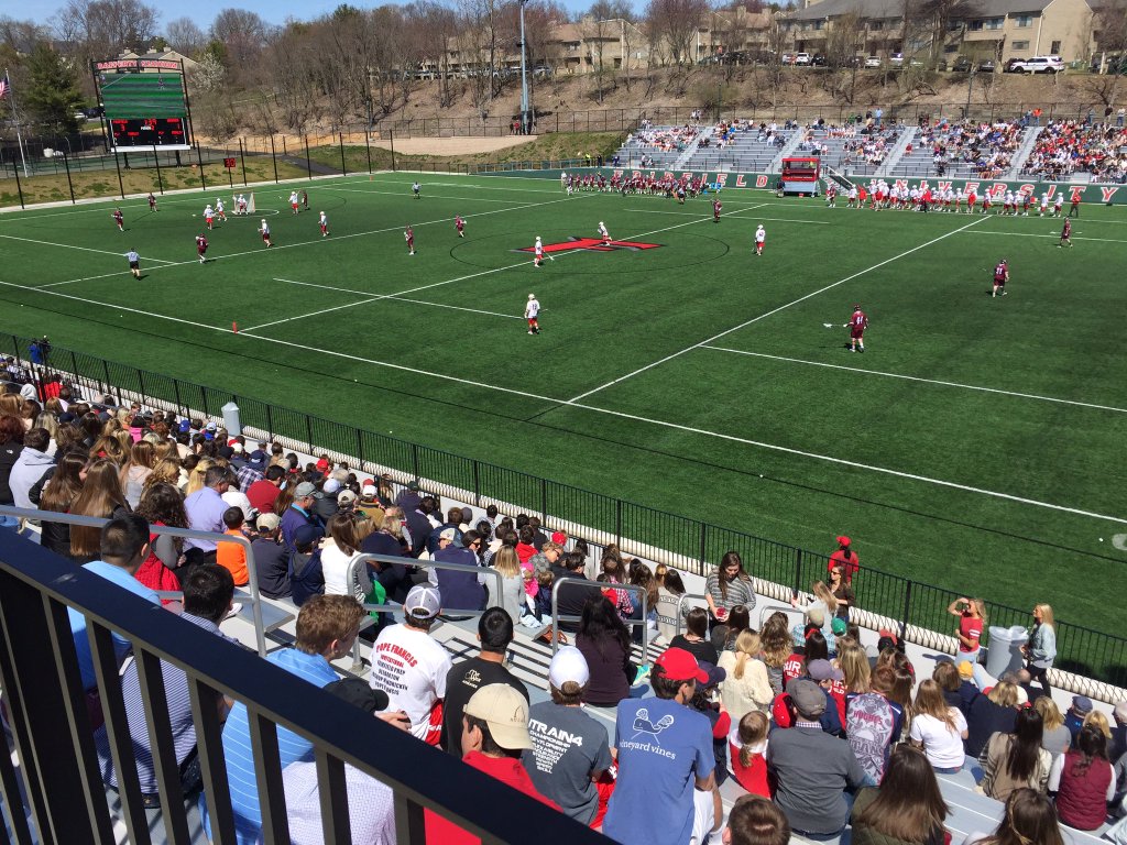 Aerial view of a lacrosse game on a green turf field with packed stadium stands.