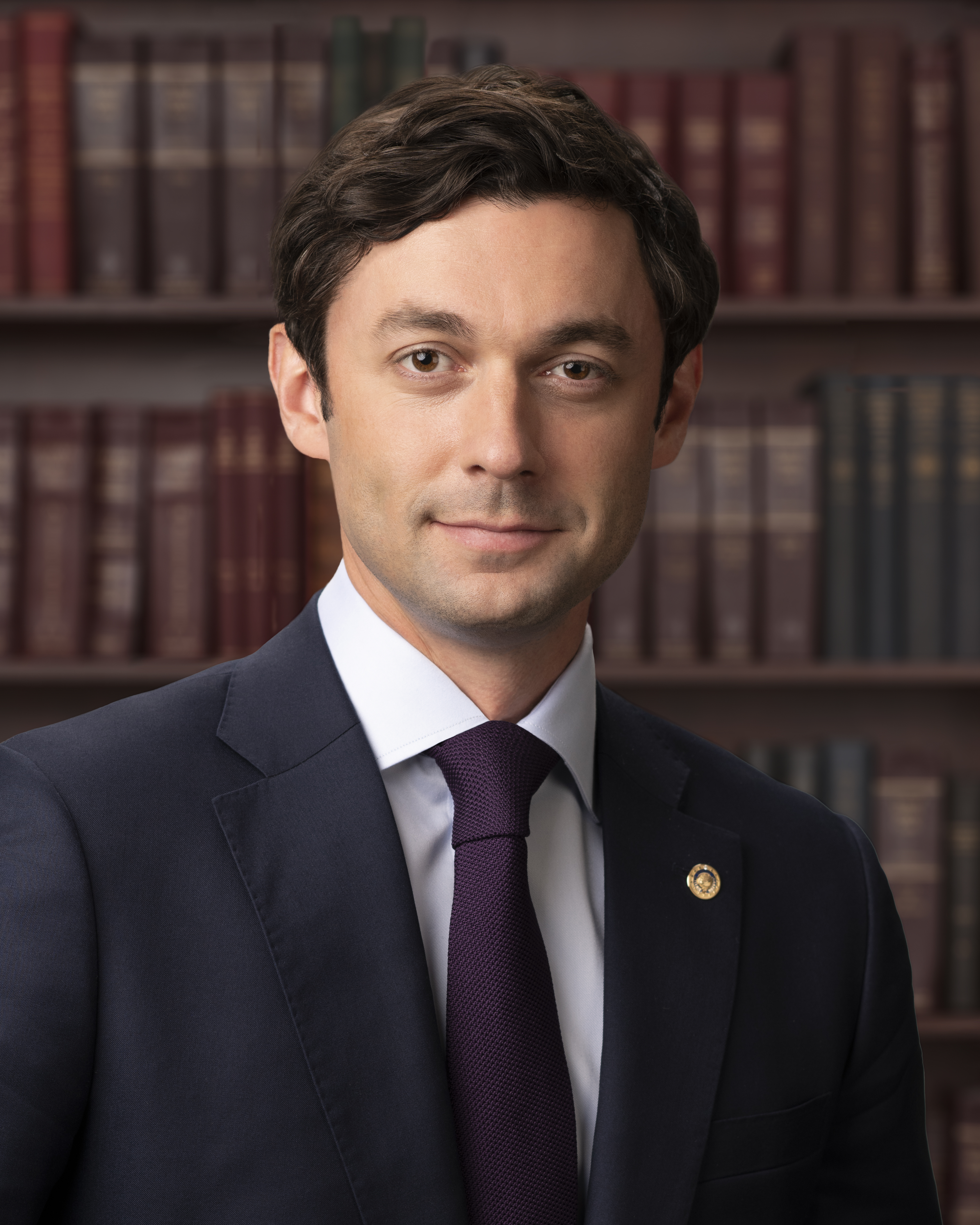 Formal portrait of a man in a dark suit and purple tie seated before bookshelves.