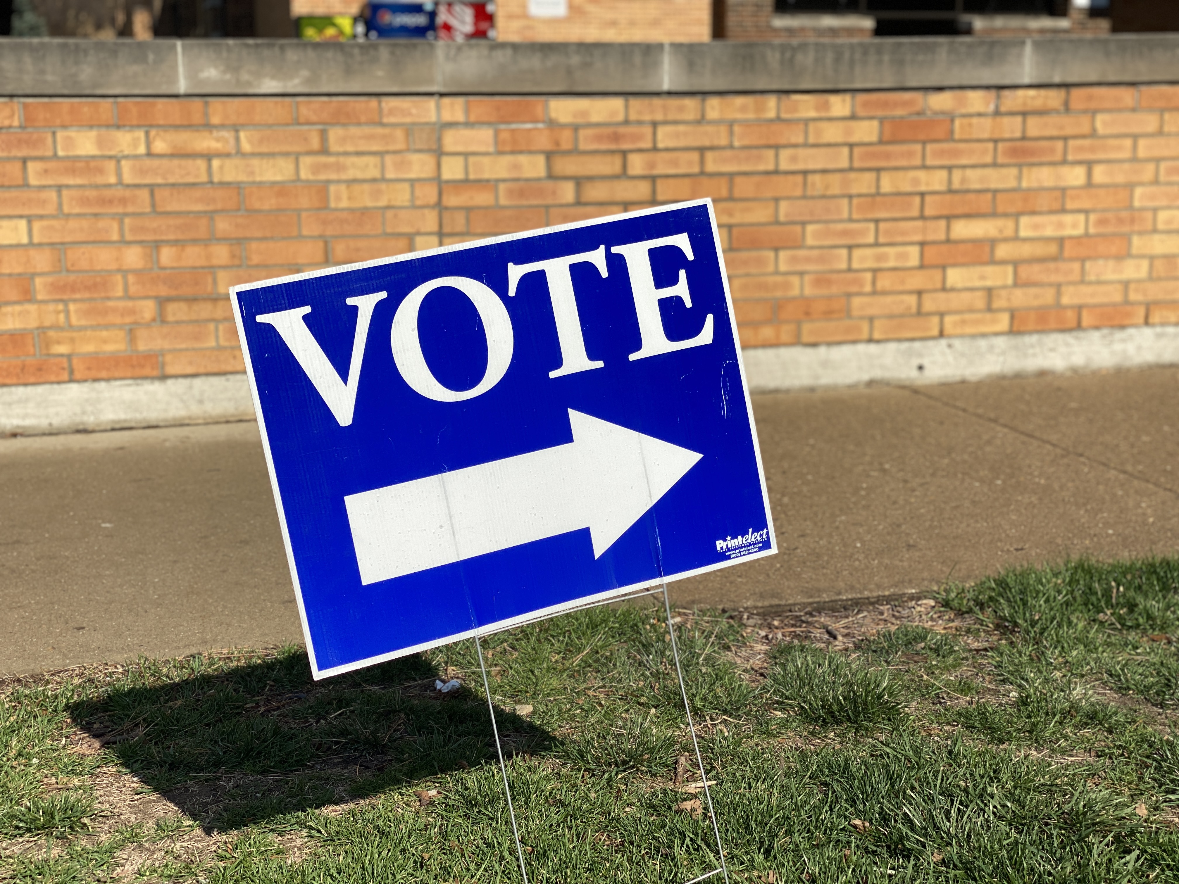 Blue yard sign reading 'VOTE' with a white arrow, staked in grass outside a brick building.