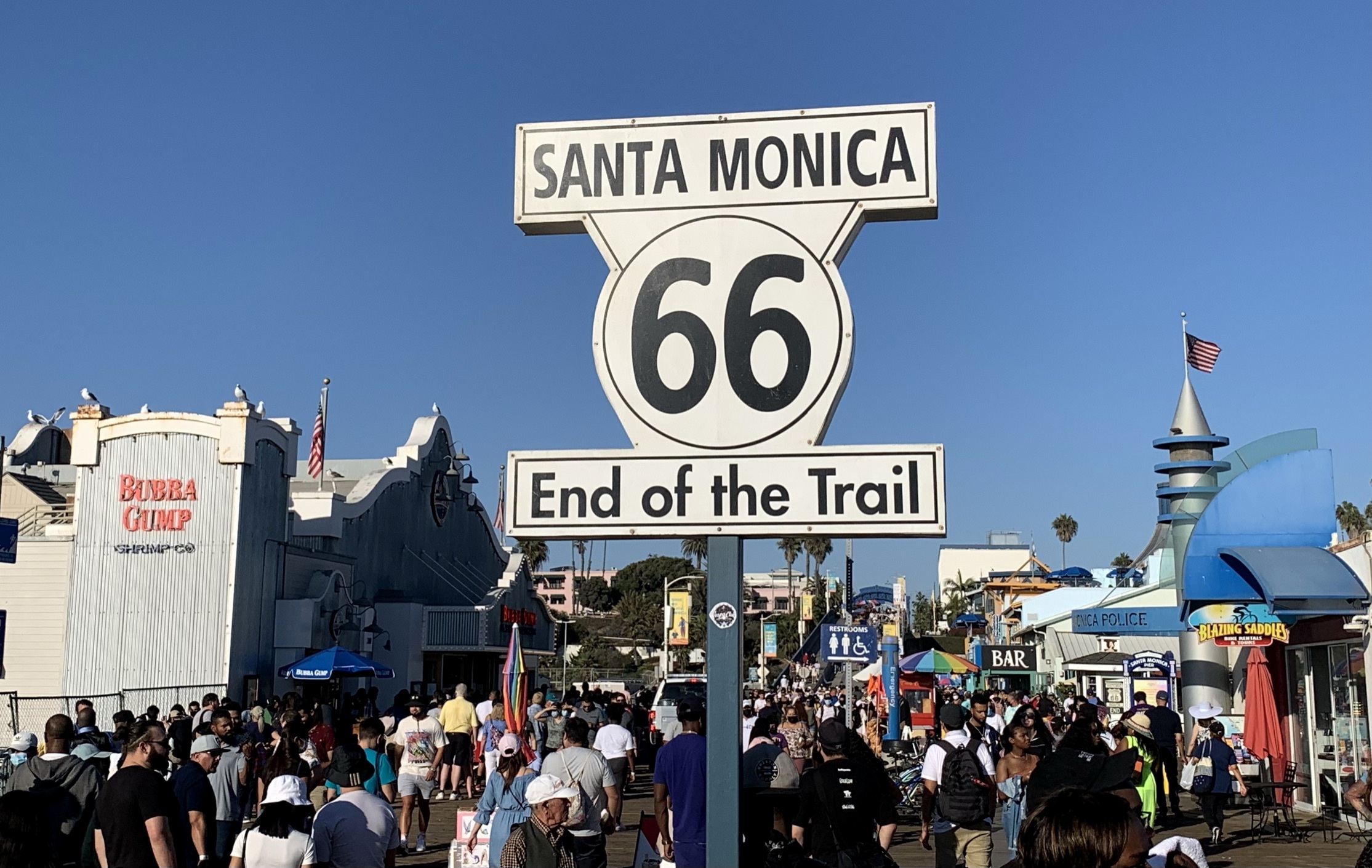 Route 66 End of the Trail sign on a pole with crowds and pier buildings behind it.