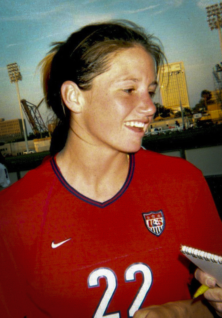 Smiling woman in a red U.S. Soccer jersey with number 22, outdoors near a stadium.