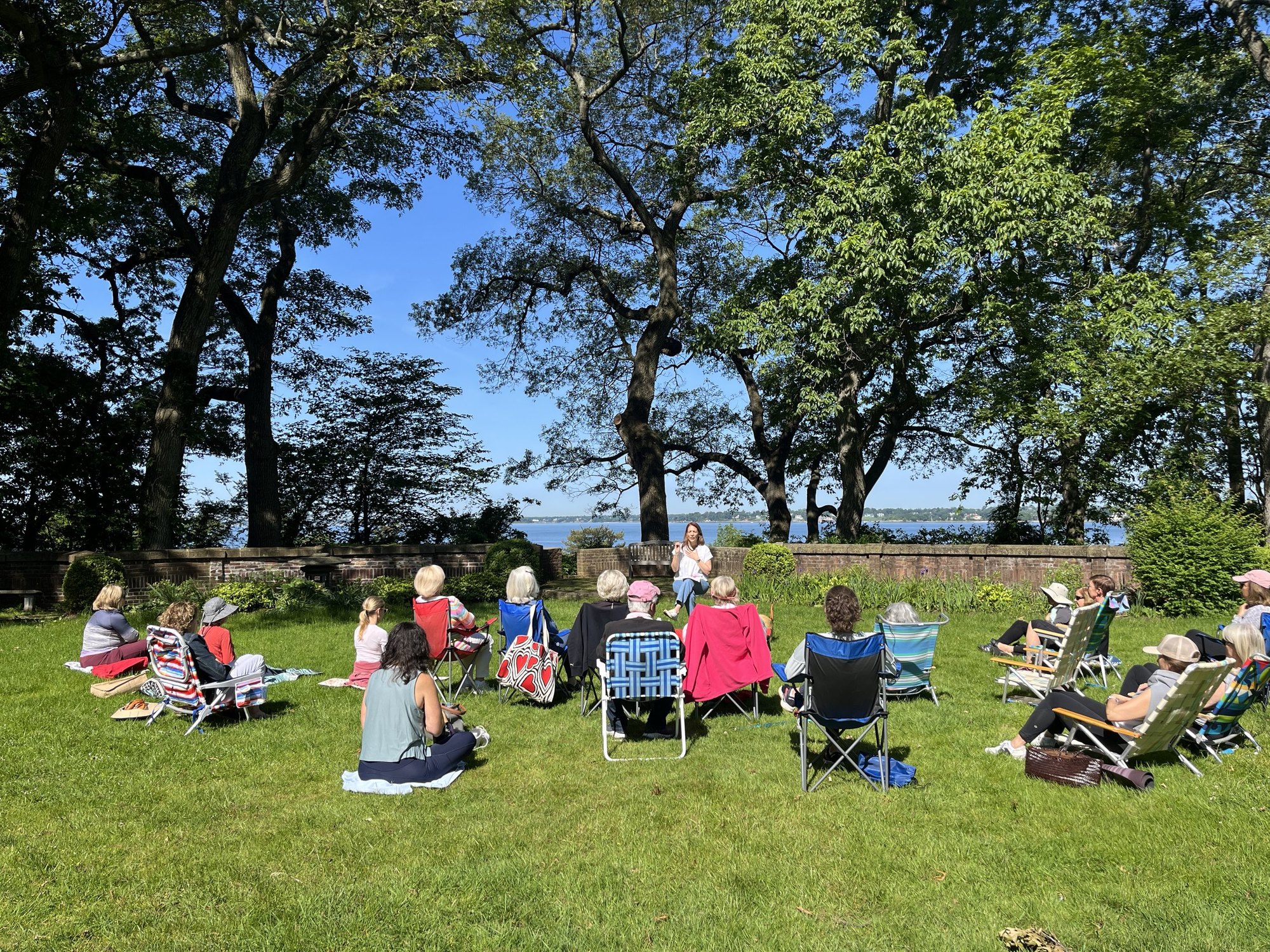 Adults seated in a semicircle of folding chairs on a grassy lawn near water.