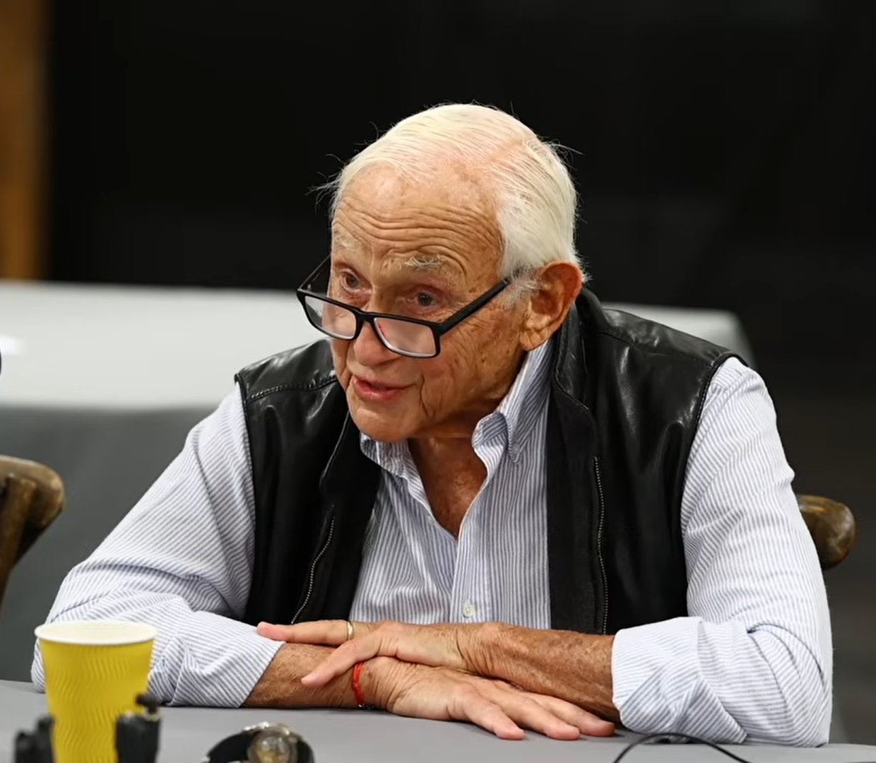 Elderly man with white hair and glasses seated at a table, arms clasped.