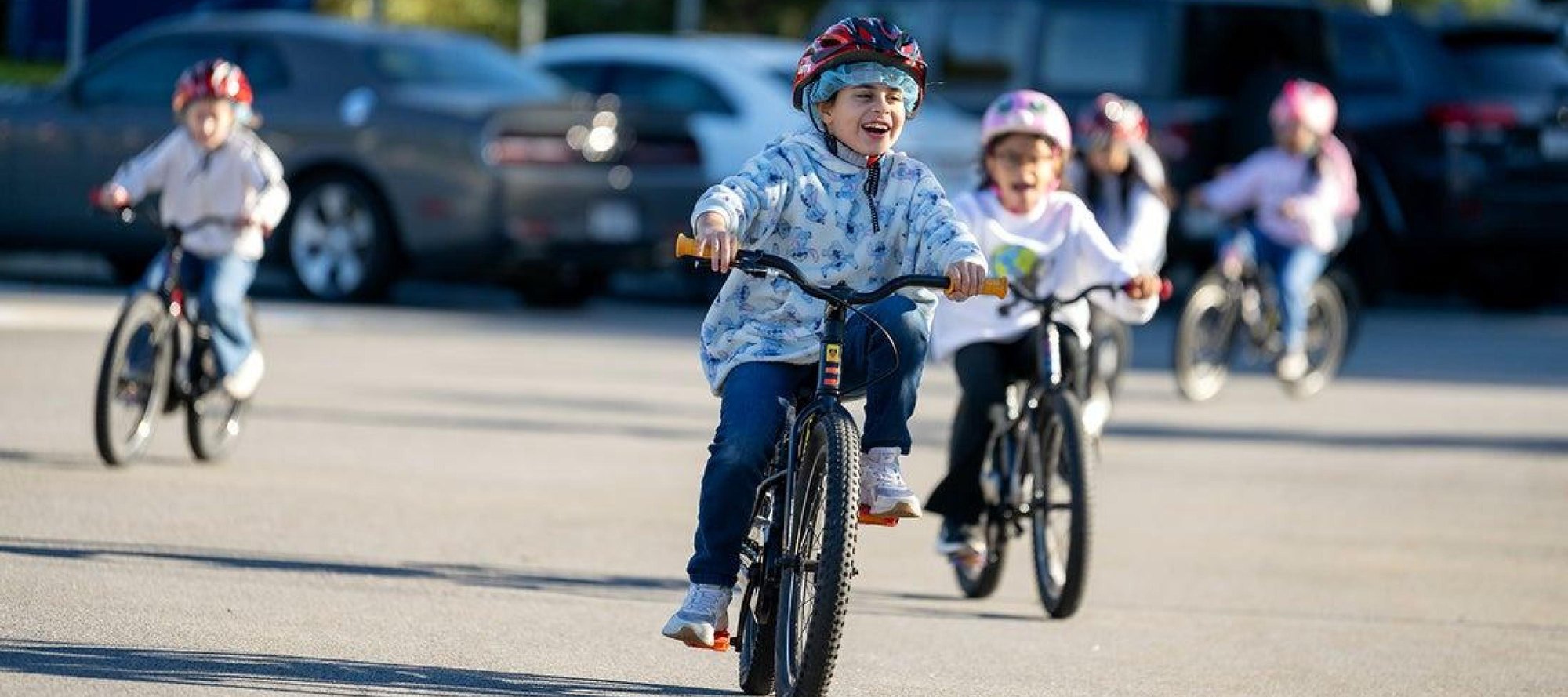 Several helmeted children smile while riding bicycles on a paved school loop.