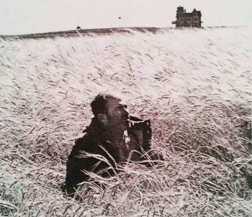 A figure crouches in a tall wheat field with a large farmhouse visible on the horizon.