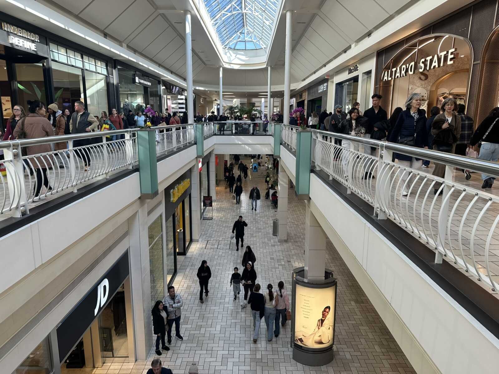 Shoppers walking through a two-level indoor shopping mall with a skylight above.