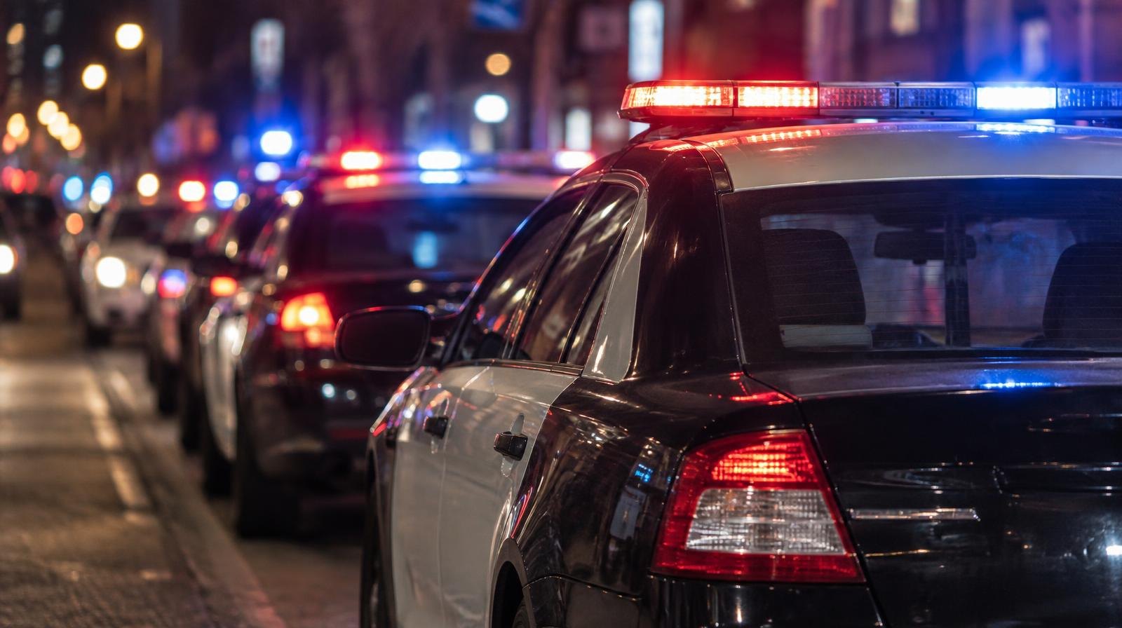 A row of police cruisers with flashing light bars parked on a commercial street at night.