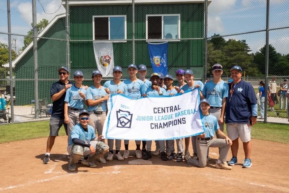 Youth baseball team in light blue uniforms poses with a championship banner on a sunny field.
