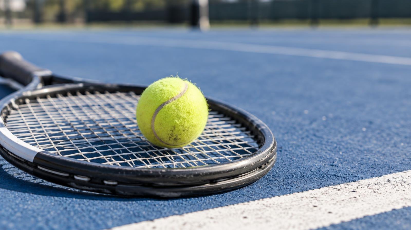 Yellow tennis ball resting on the strings of a tennis racket on a blue hard court.