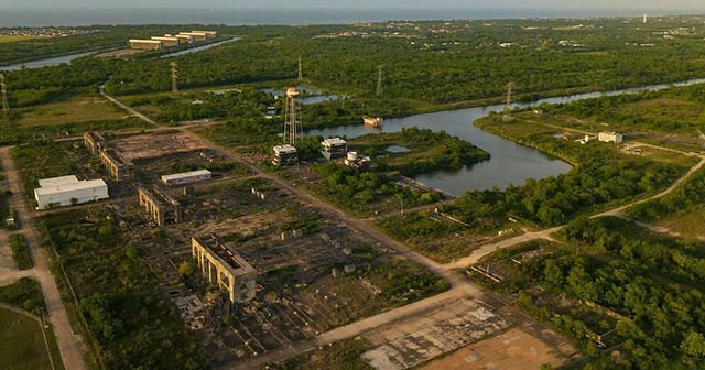 Aerial view of a ruined industrial site with transmission towers and a bay visible in the background.