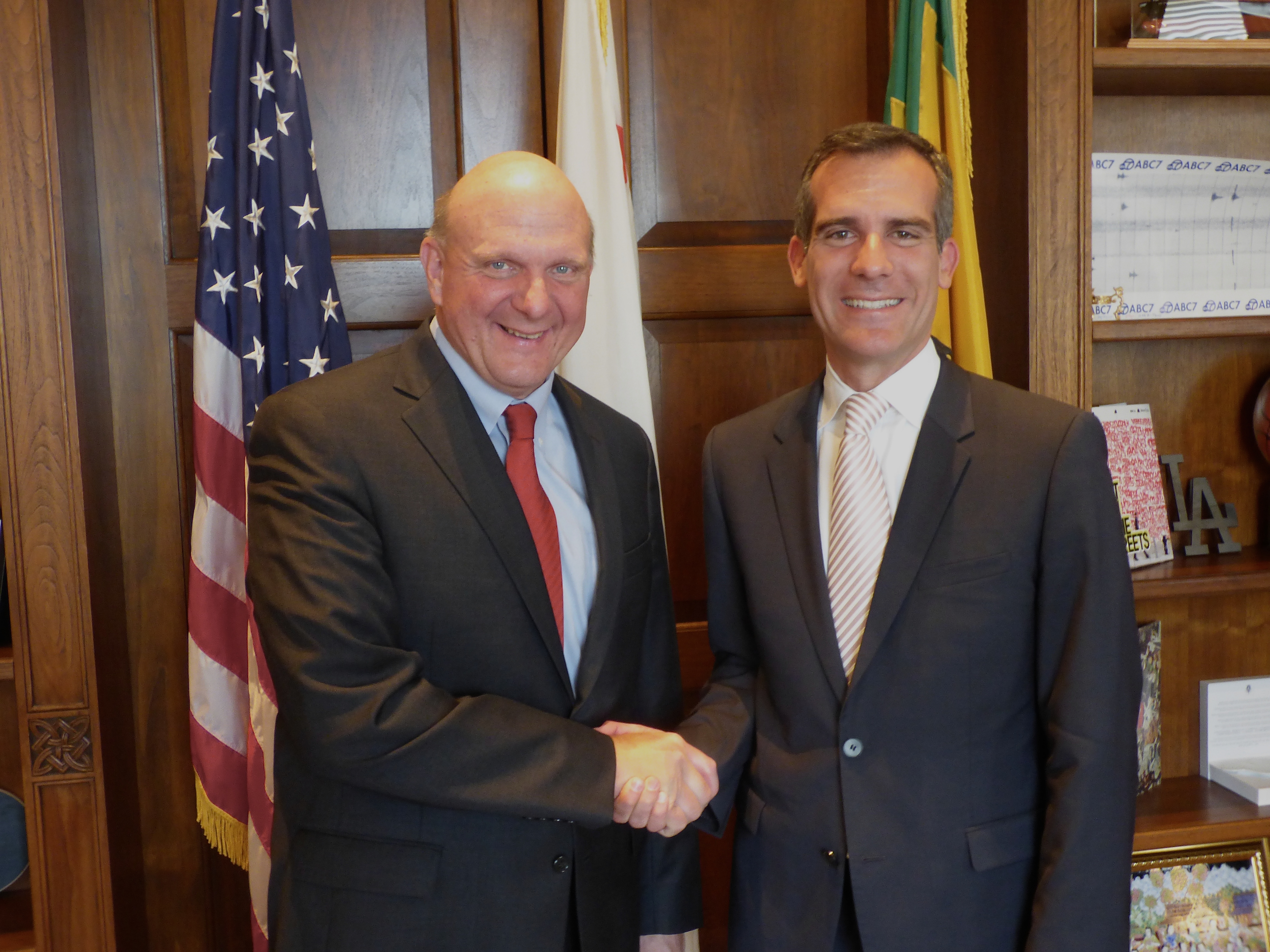 Two men in suits shaking hands in a formal office with flags in the background.
