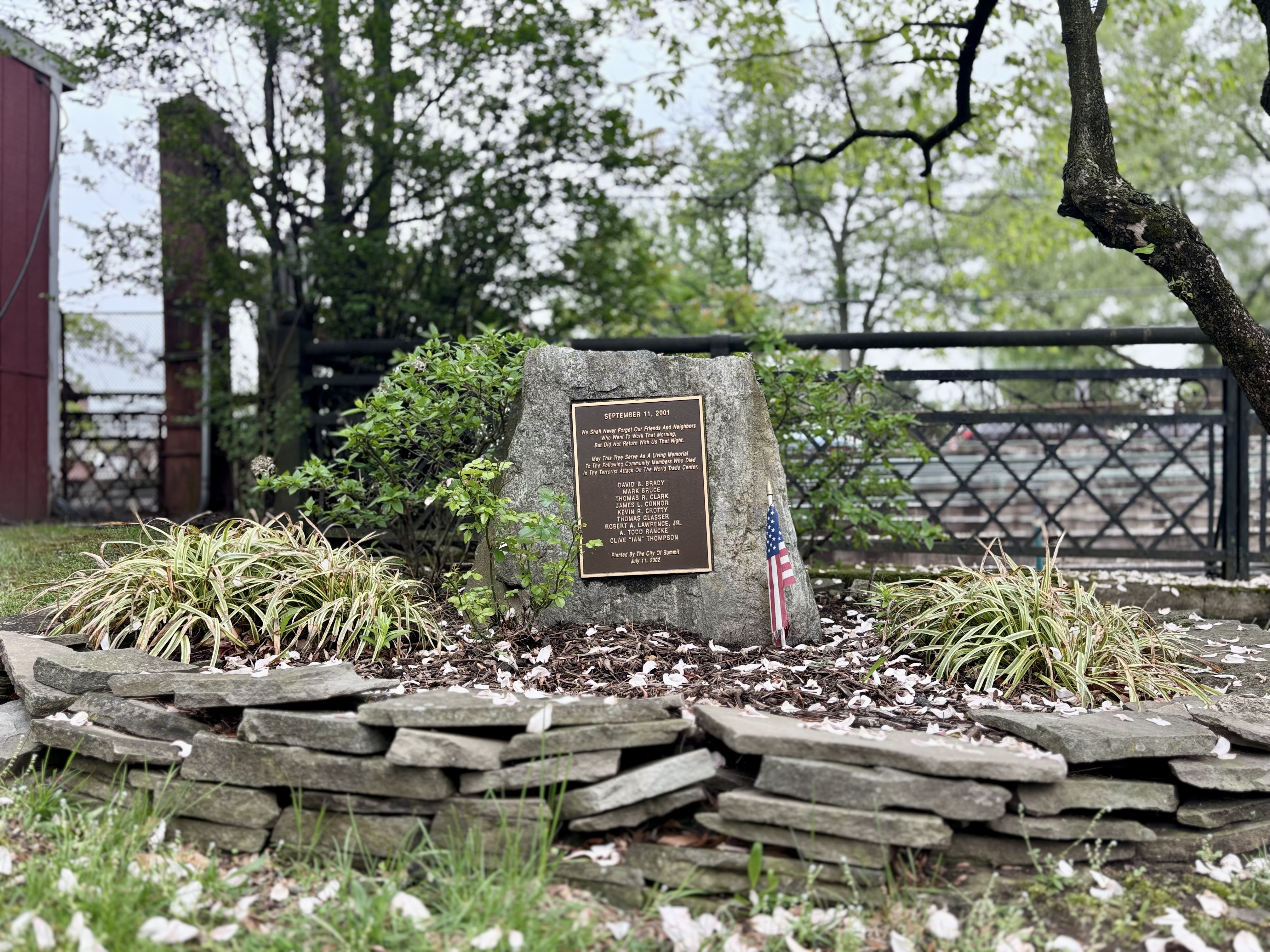 Stone memorial monument with bronze plaque and American flag near a fence outdoors.