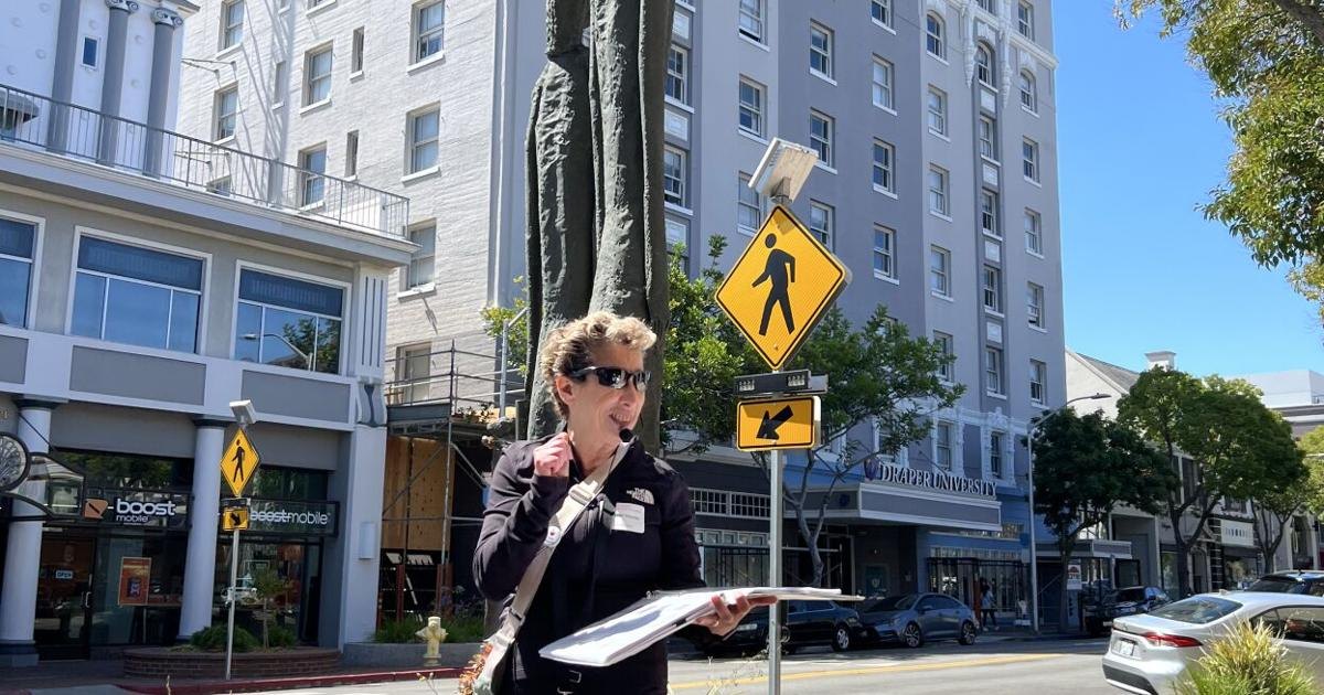 Woman in sunglasses and dark blazer holding papers on a downtown sidewalk with multi-story buildings behind her.