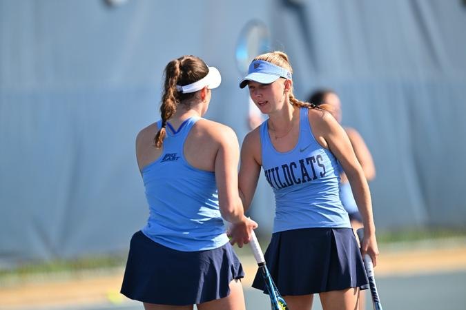 Two female tennis players in light blue uniforms fist-bumping on a tennis court.