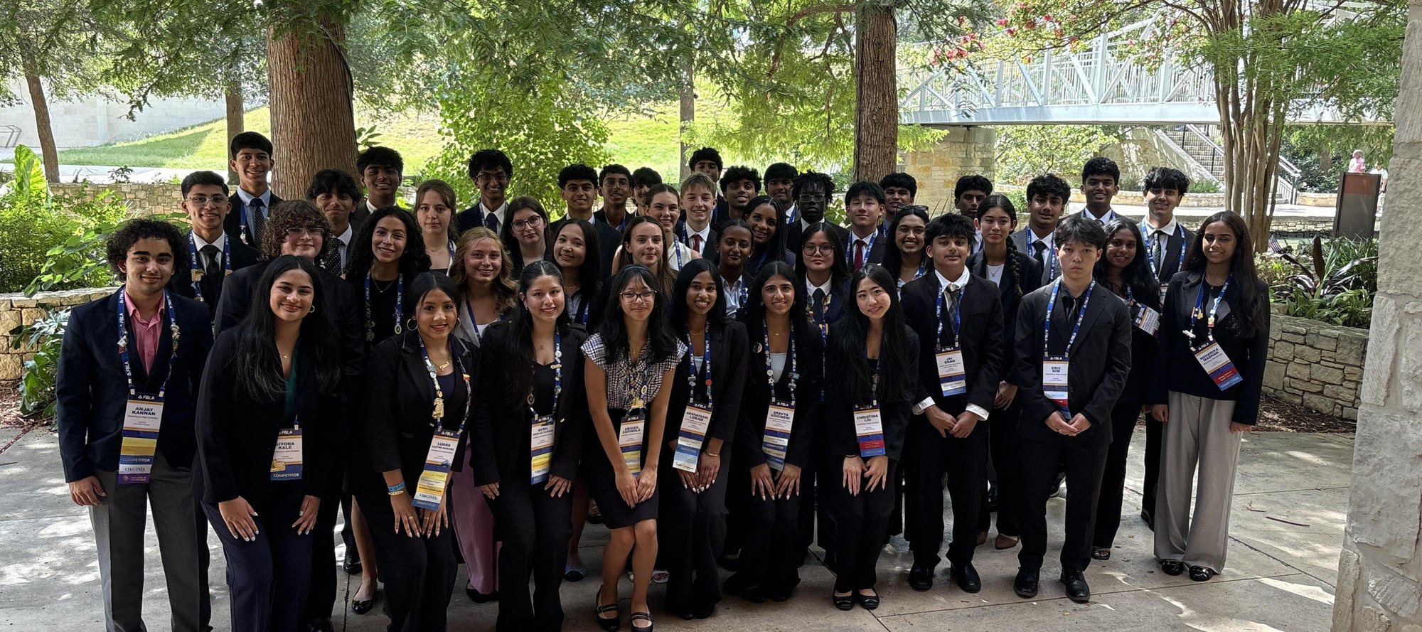 Large group of students in dark blazers and conference lanyards posing outdoors at a landscaped venue.