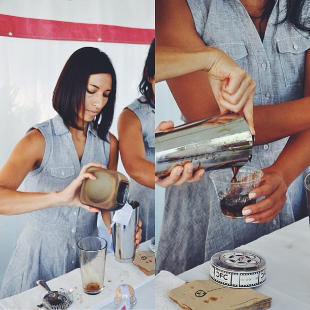 Woman pouring a drink from a metal cocktail shaker into a glass at an outdoor event.
