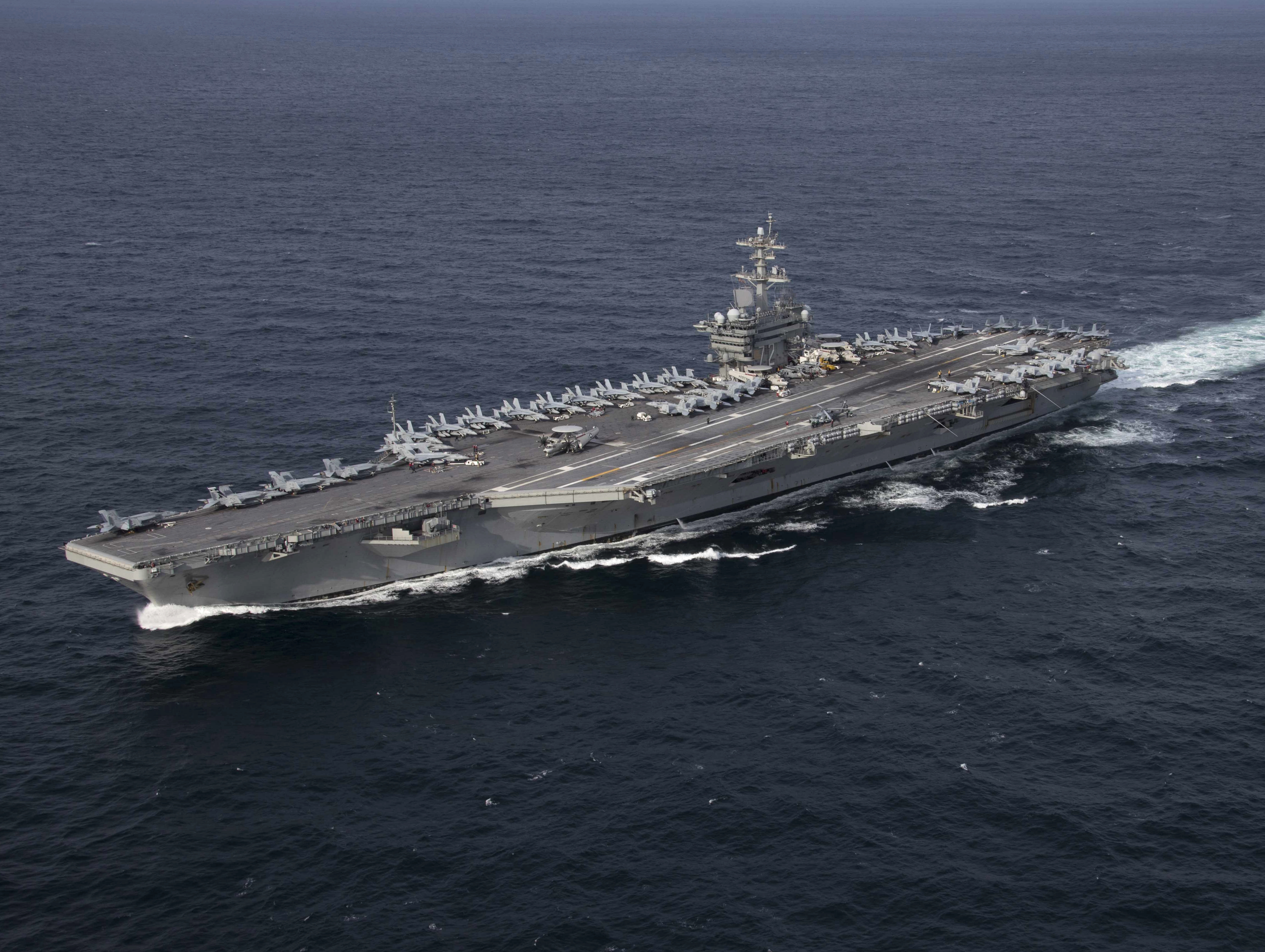 Aircraft carrier underway at sea with aircraft on the flight deck and a white wake trailing behind.