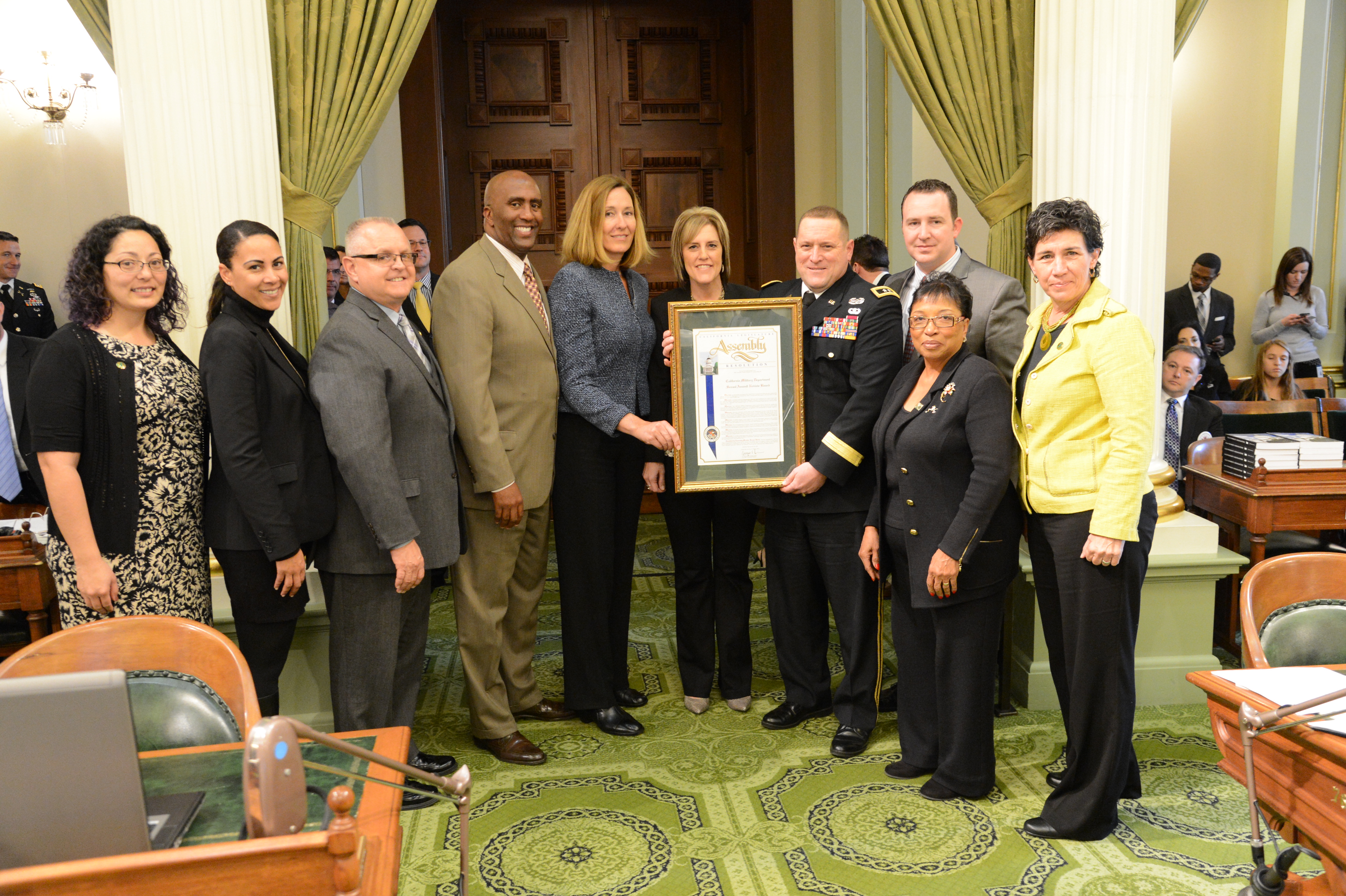Group of people in formal attire on a legislative chamber floor, two holding a framed document.